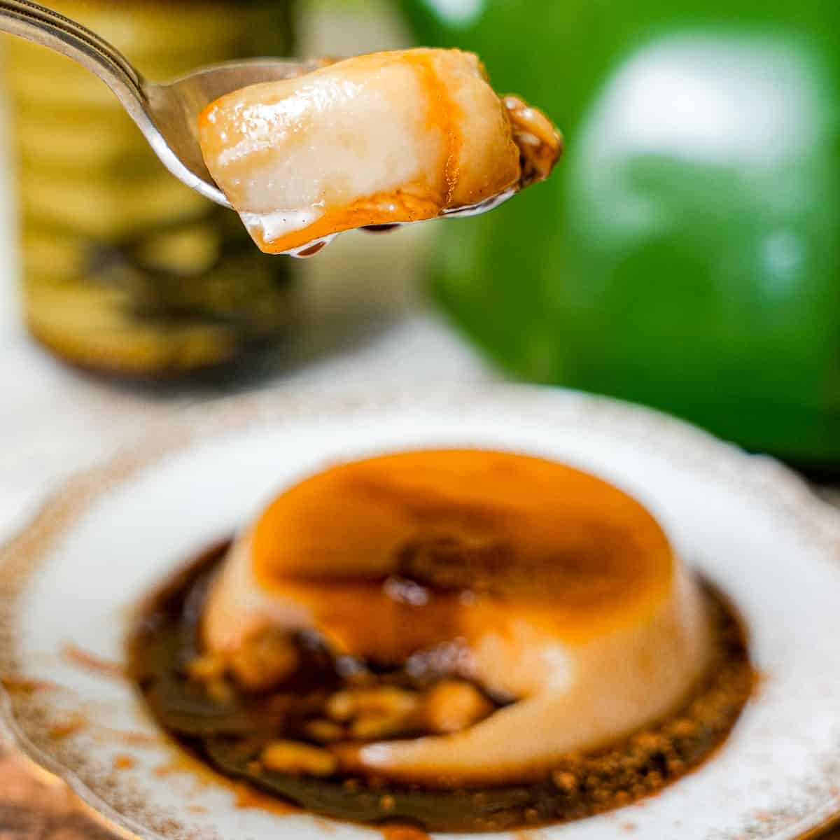 A spoonful of banh flan dripping with caramel is held over a plate with flan. A tea pot and glass can be seen in the background.
