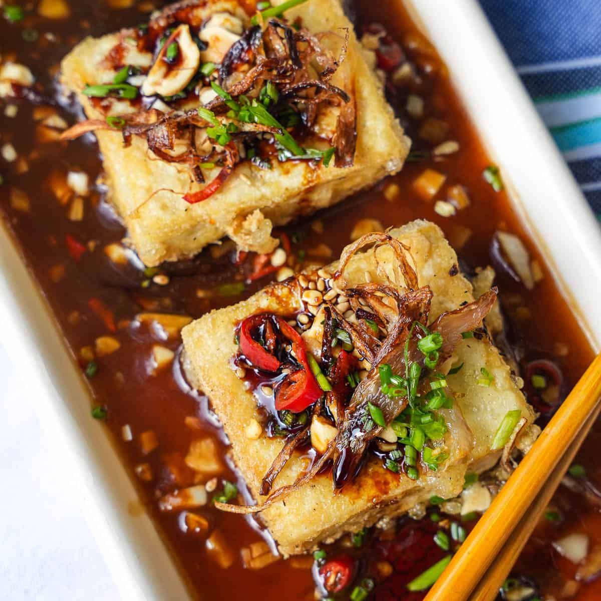 Two squares of tofu in sauce with chopsticks.
