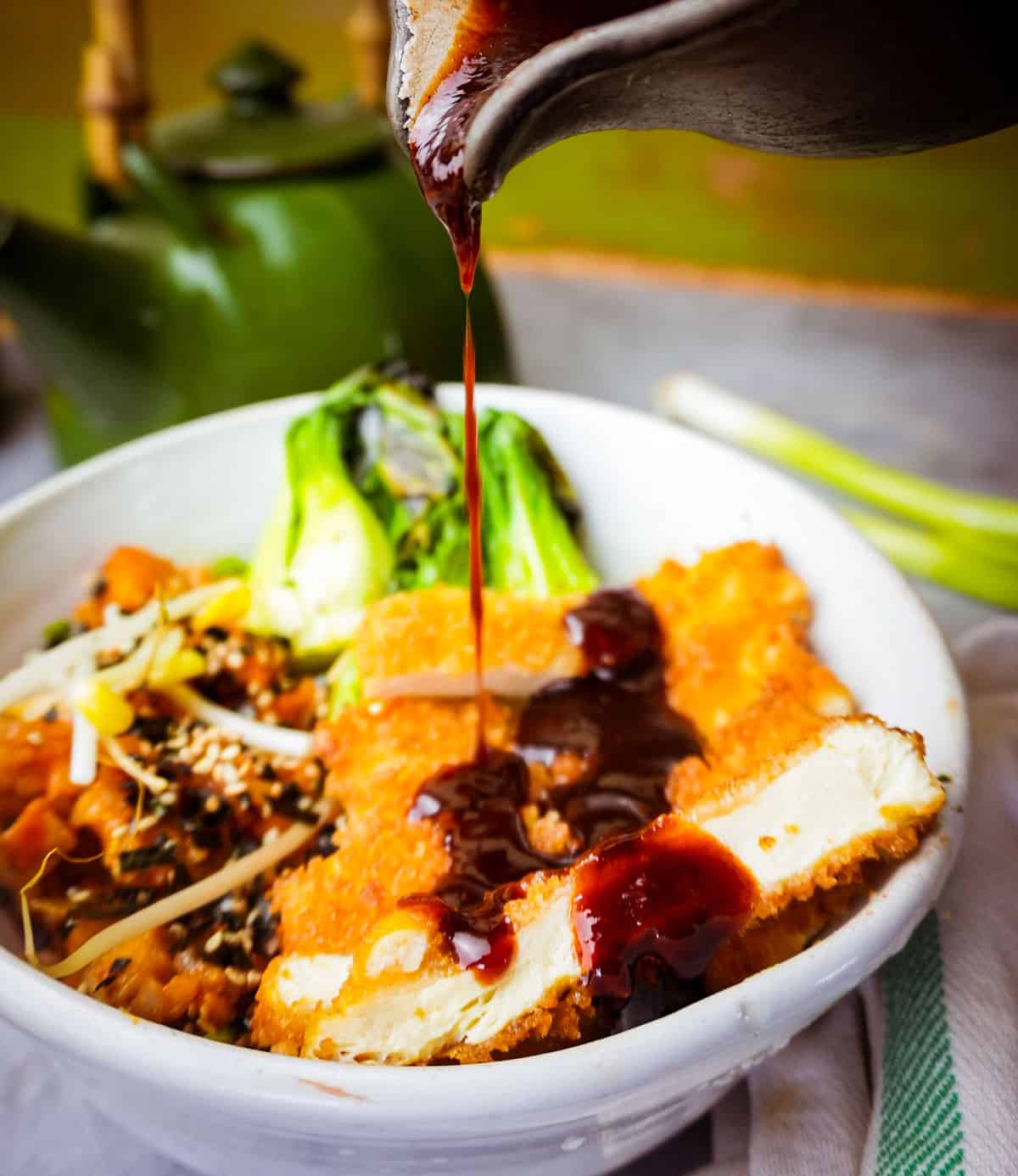 Tofu katsu in a white bowl getting tonkatsu sauce drizzled on it from a pitcher. scallions and tea kettle behind it.