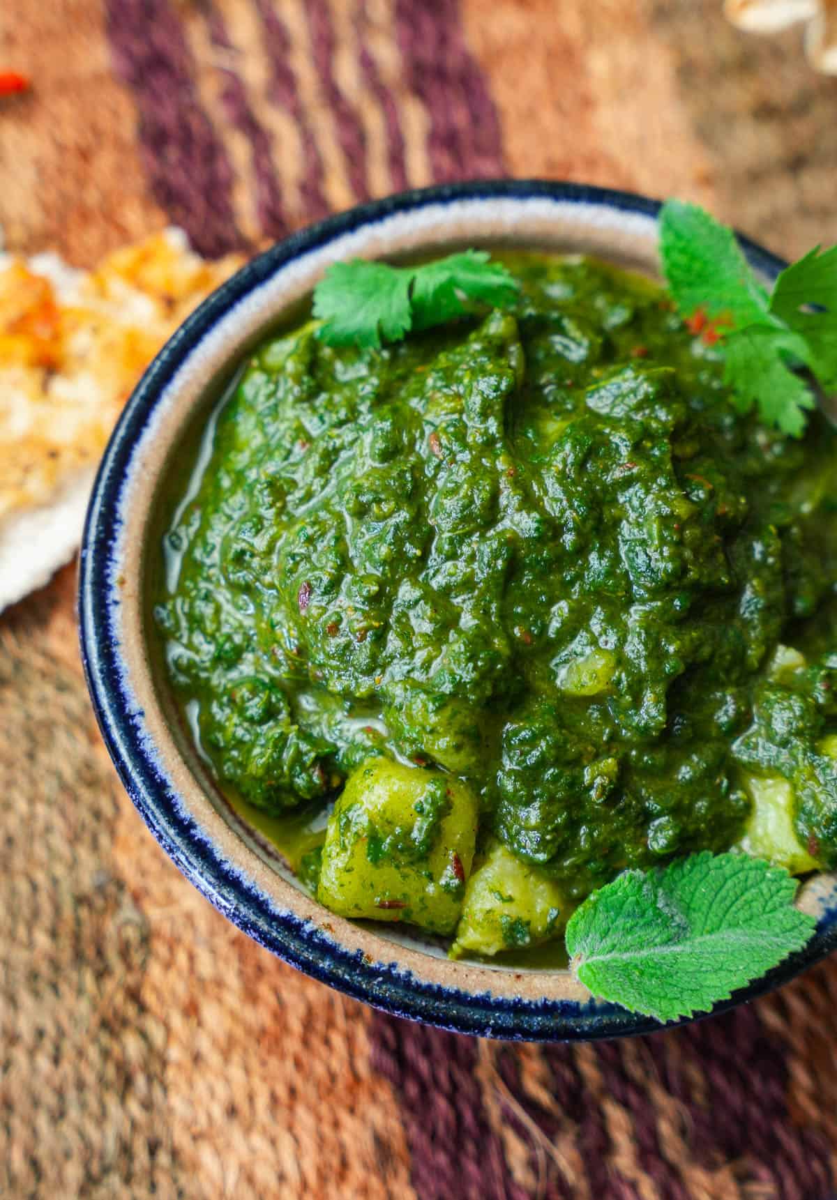 Saag aloo garnished with cilantro and mint leaves in a blue rimmed ceramic bowl on a woven mat with a piece of naan.