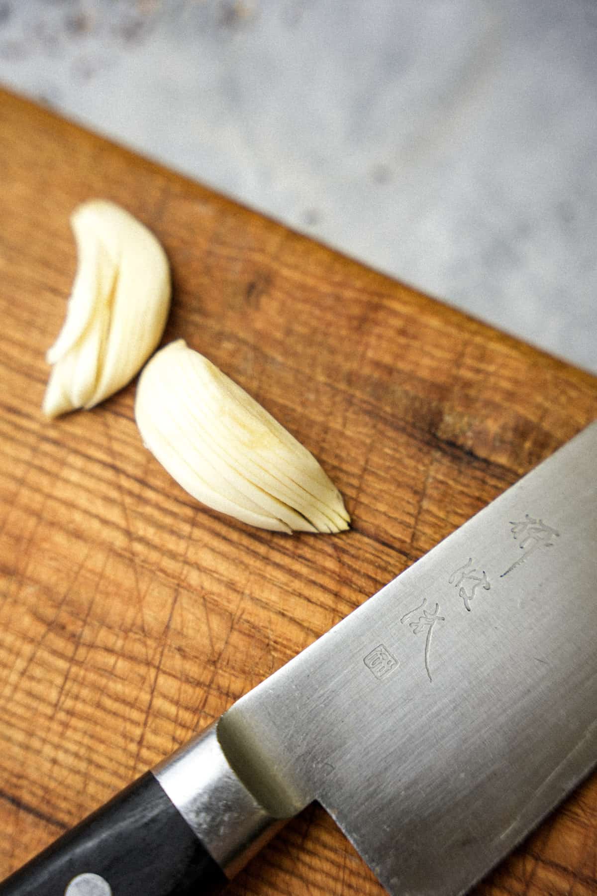 Thinly sliced garlic cloves next to a Japanese knife on a wooden cutting board.