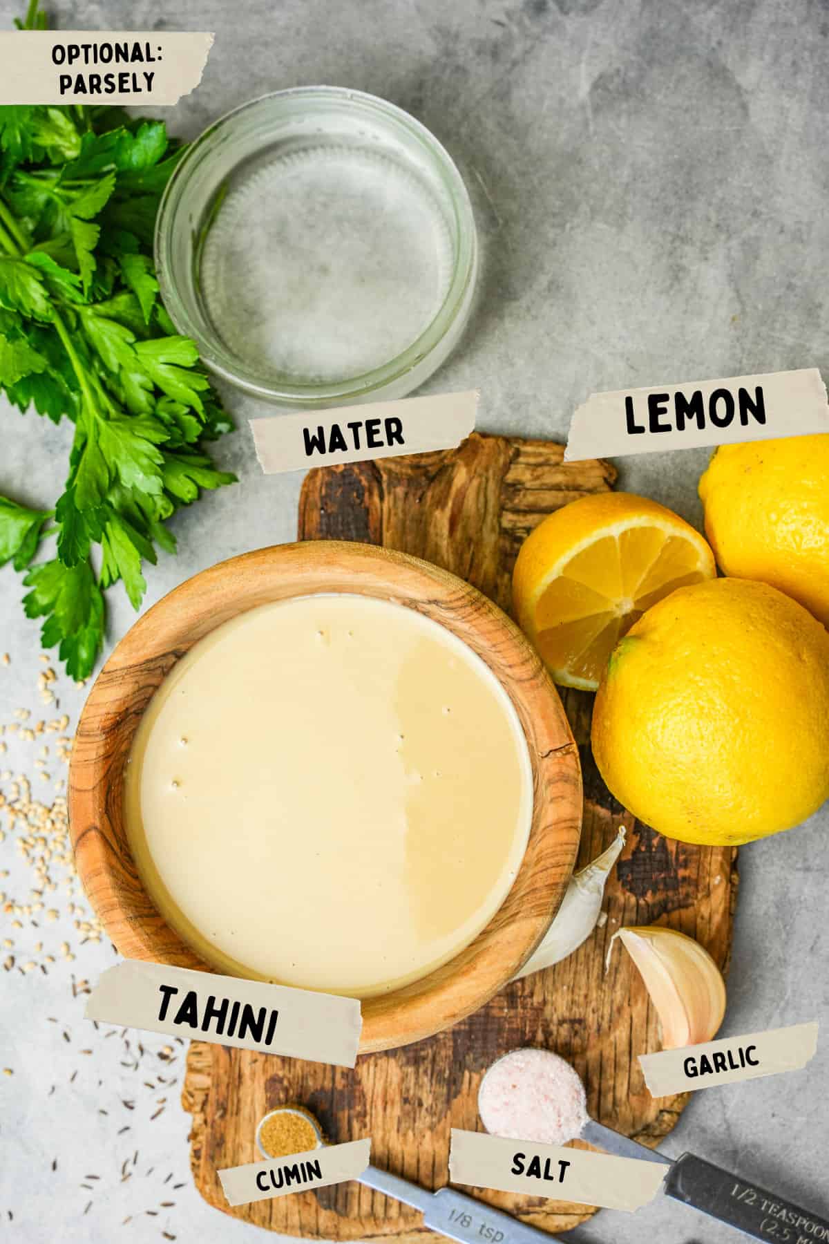 Ingredients for tahini sauce are laid out on a wooden slab on a stone countertop.