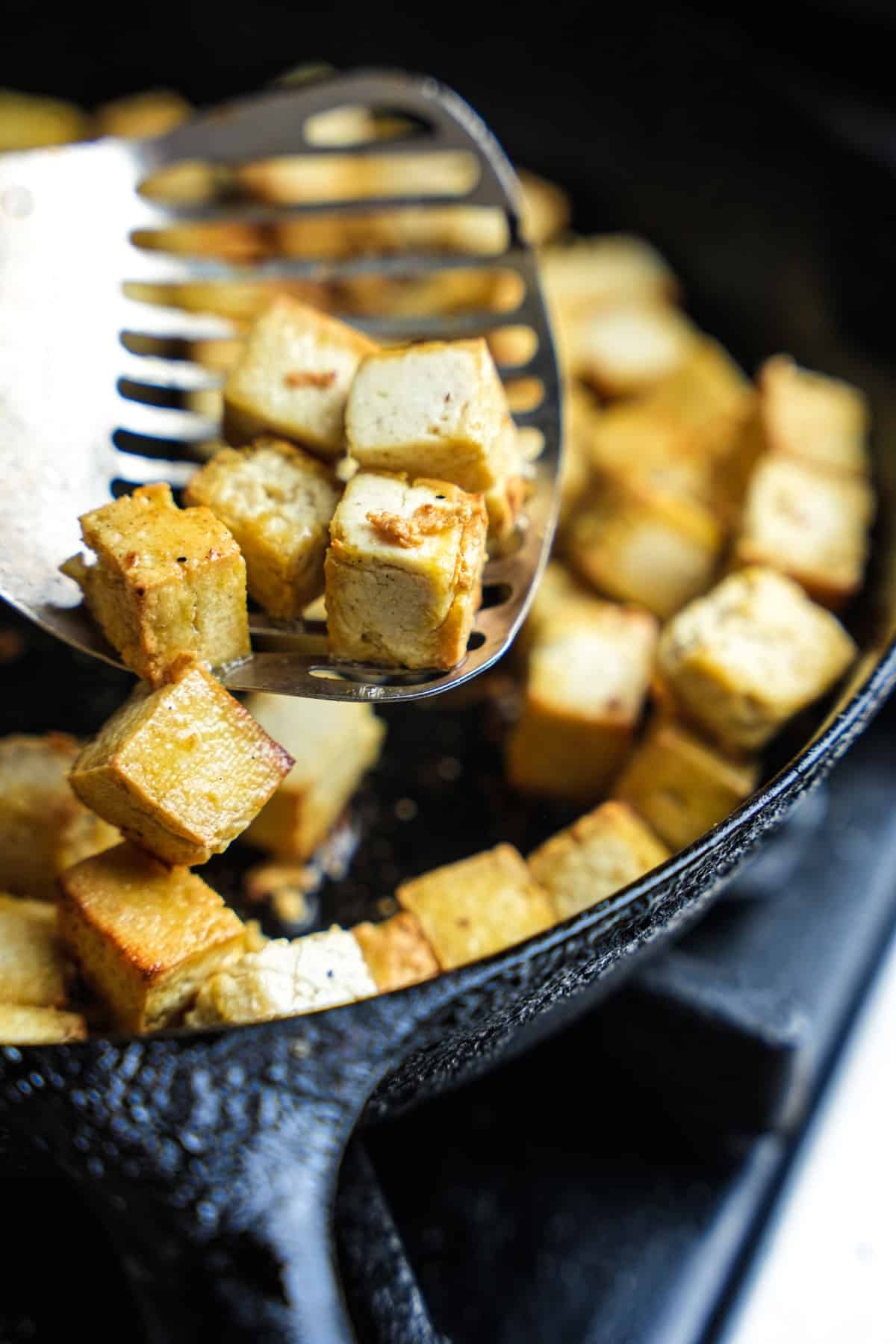 Frying tofu being flipped with a spider in an iron skillet.