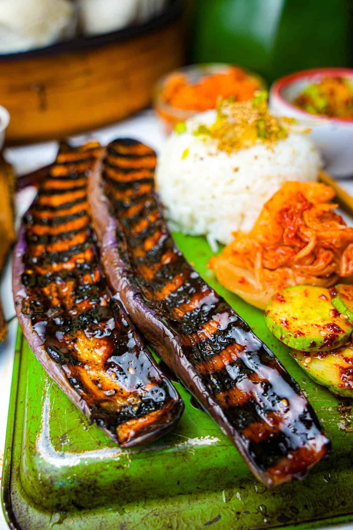 Grilled eggplant, with sides of oi muchim, kimchi, and white rice on a green metal tray.