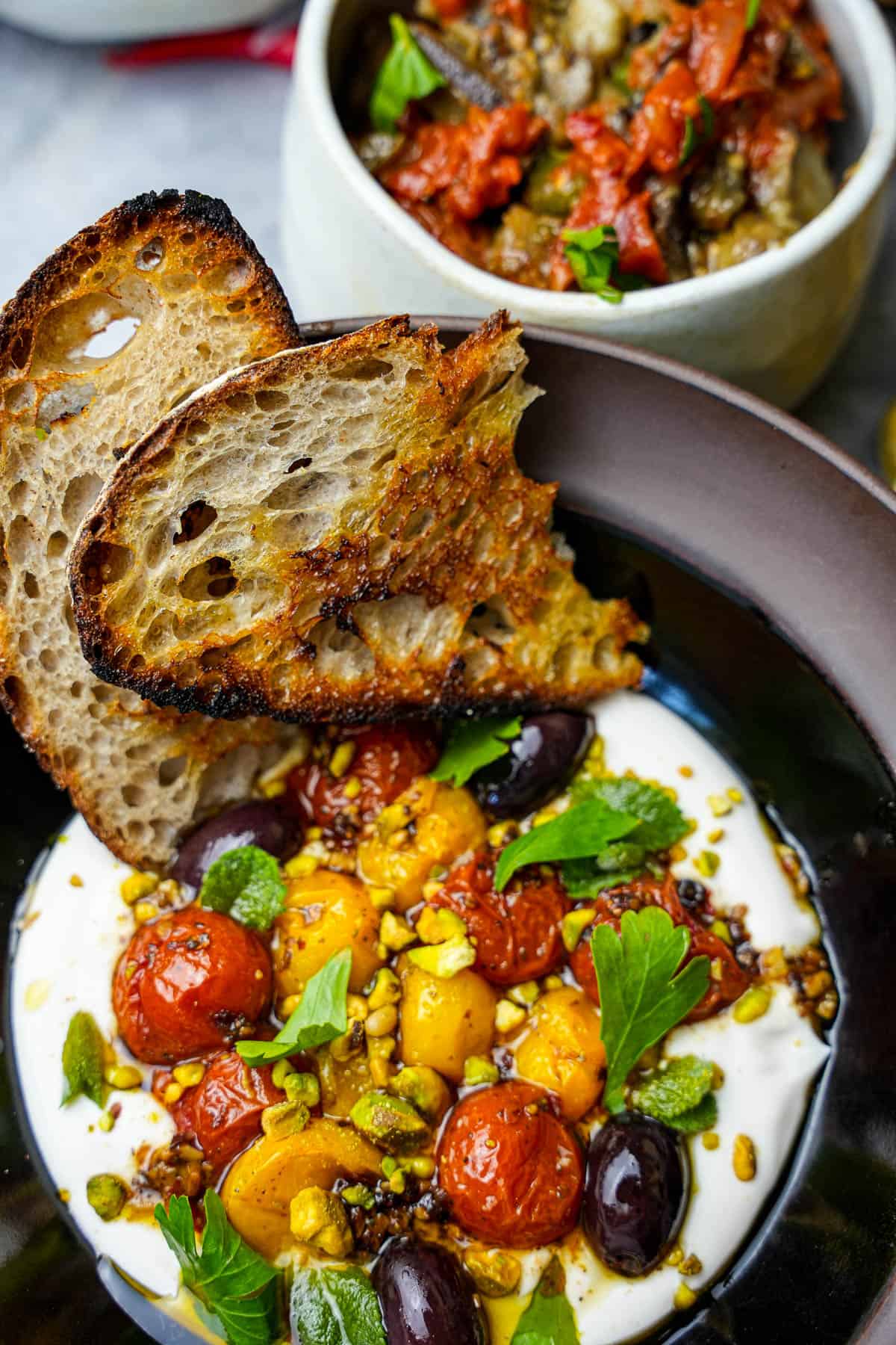 Labneh in a black bowl topped with roasted tomatoes, herbs and pistachios. Grilled bread, and Turkish shakshuka on the side.
