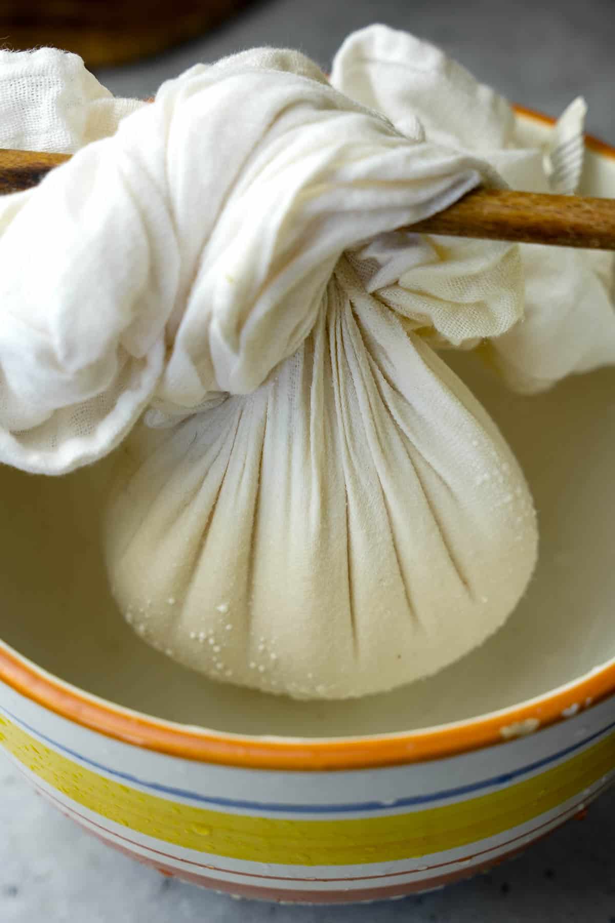 A portion of labneh draining into a bowl. The cheesecloth package is suspended in the bowl from a wooden spoon it is tied around.