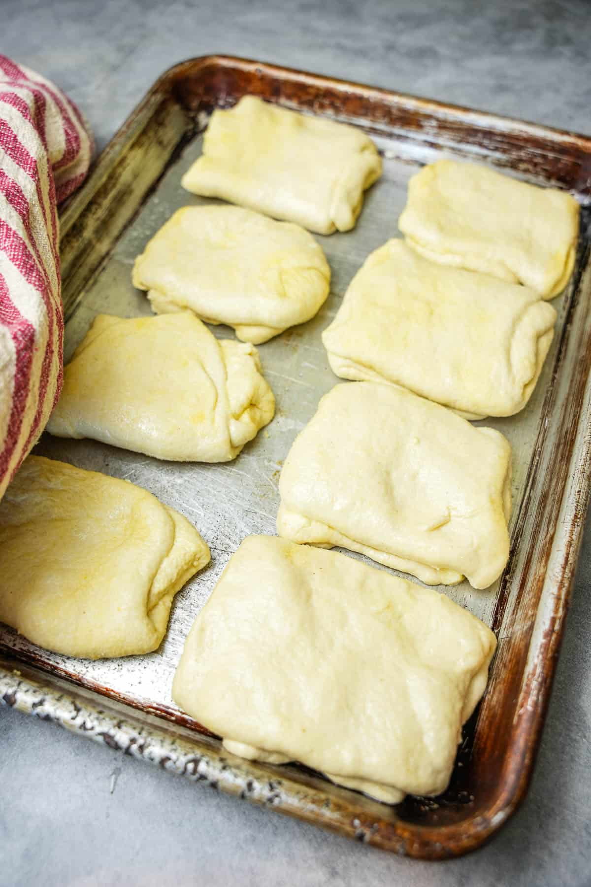 A metal sheet pan holding 8 prepared, layered portions of dough waiting to be cooked.