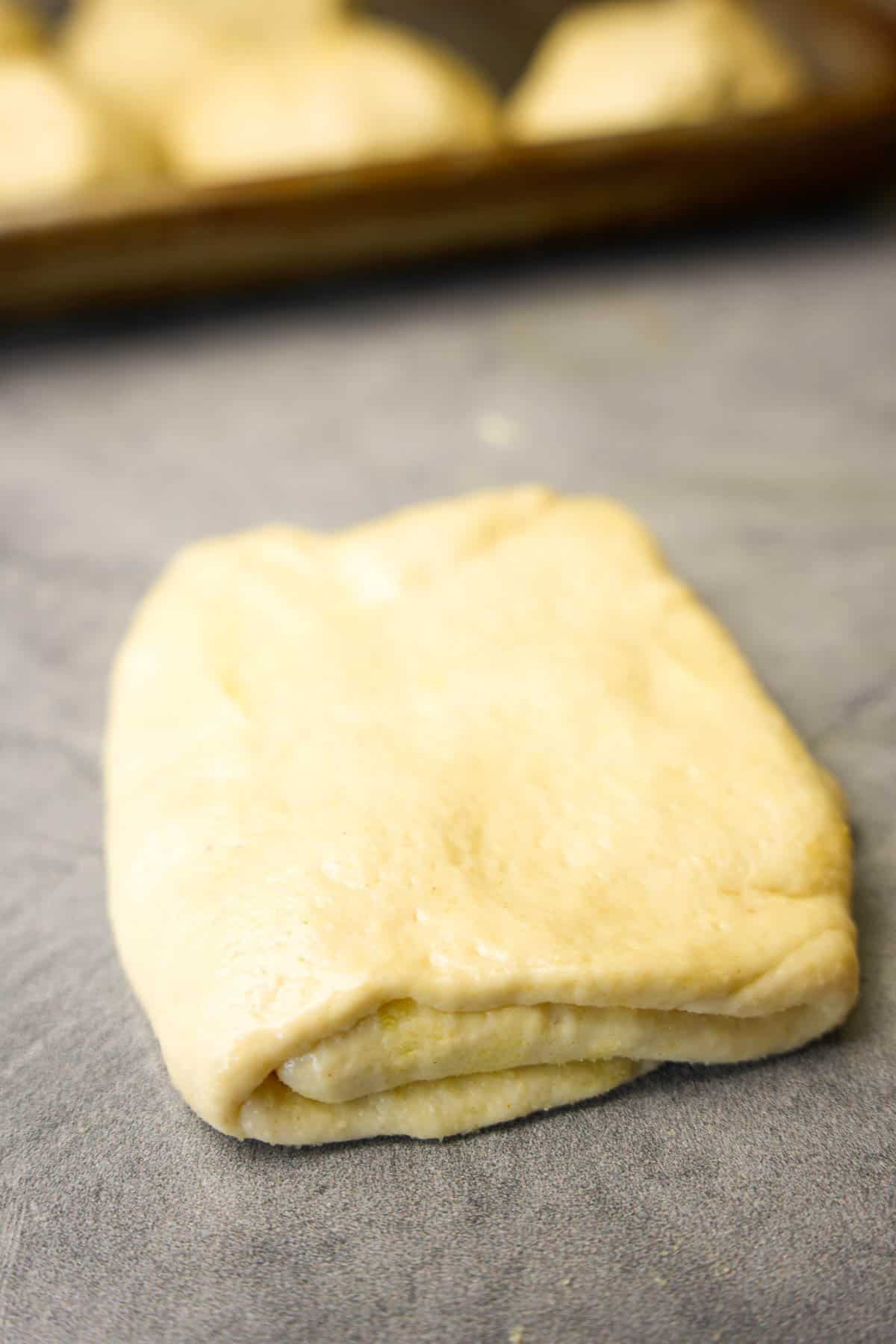 A close up showing a single formed portion of msemen dough waiting to be cooked. A tray in the background shows dough waiting to be formed.
