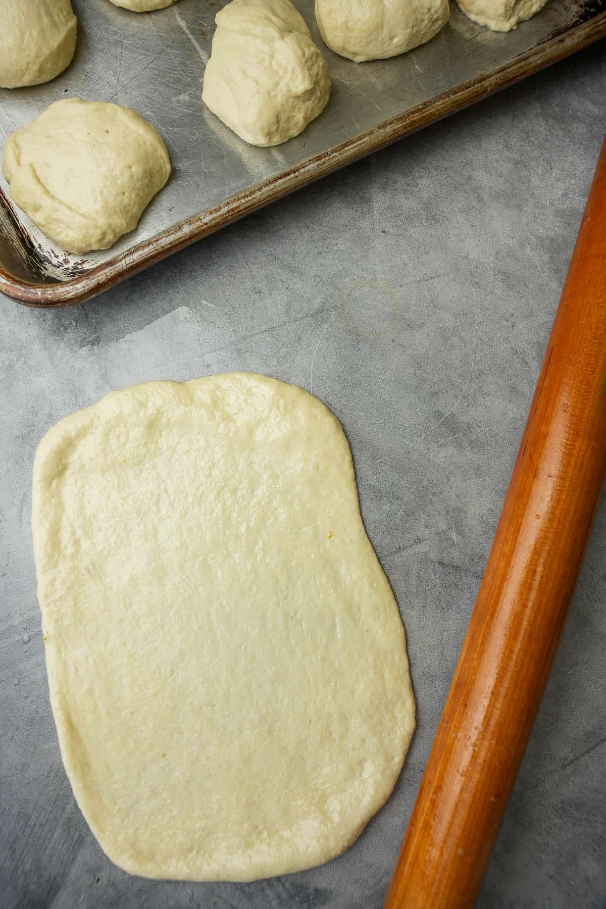 A portion of dough is rolled out to form a thin rectangle. A rolling pin and tray of dough balls are also on the table