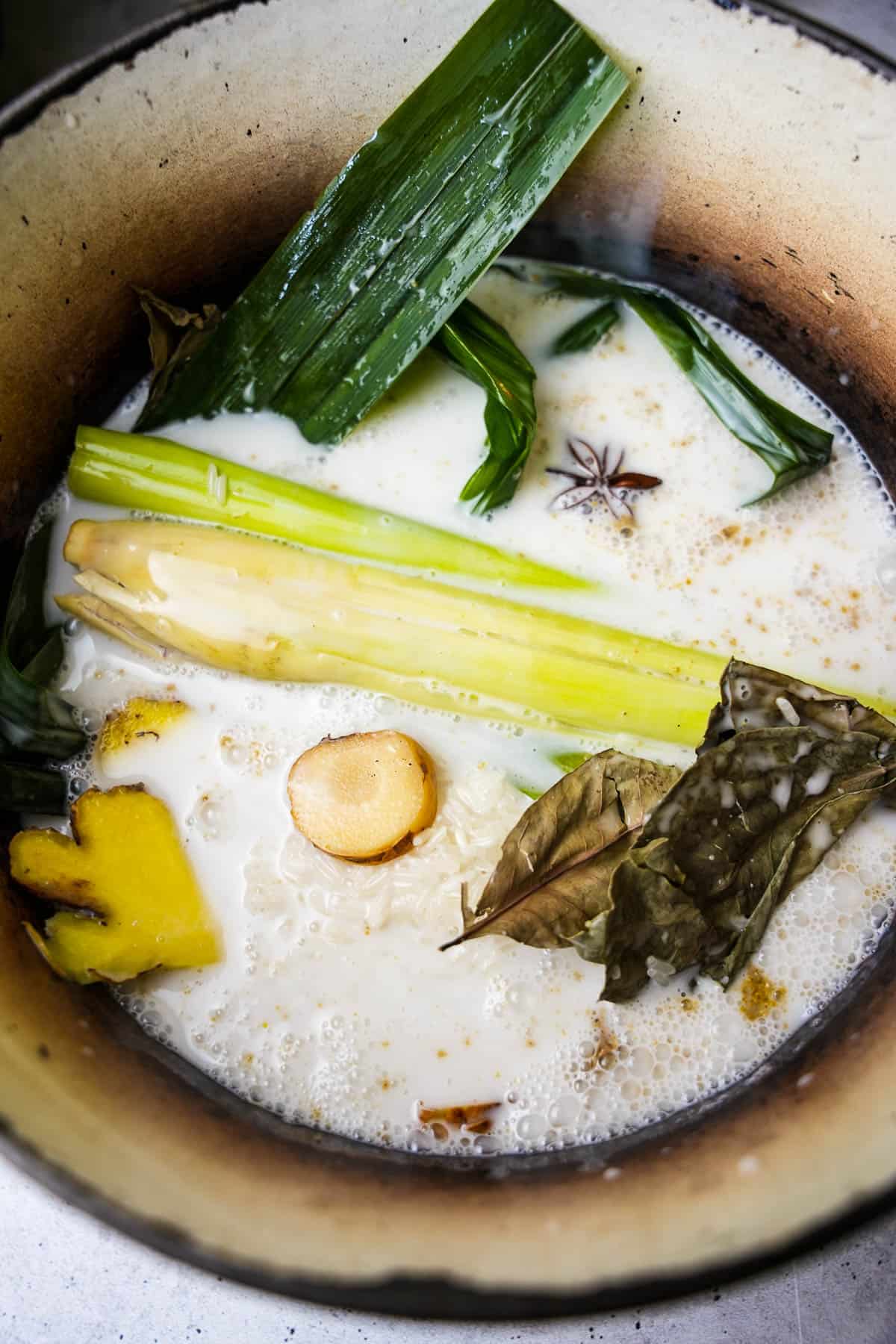 A ceramic pot containing all of the rice ingredients. You can see the Indonesian bay leaves, ginger, galangal, pandan, lemongrass, and star anise floating in the coconut milk.