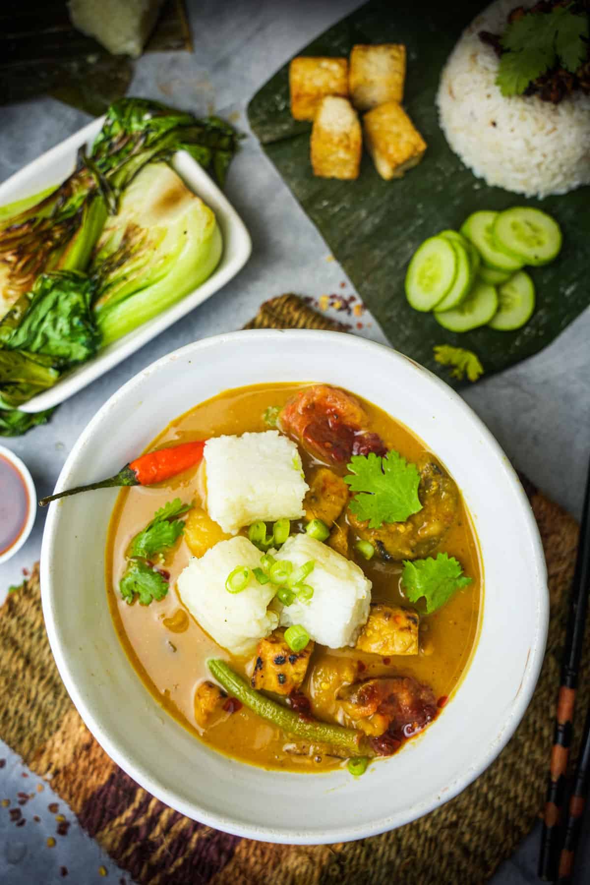 A small white bowl filled with sayur lodeh topped with lontong, scallions, sambal oelek and fresh cilantro leaves. Grilled bok choy and Indonesian coconut rice in the background.
