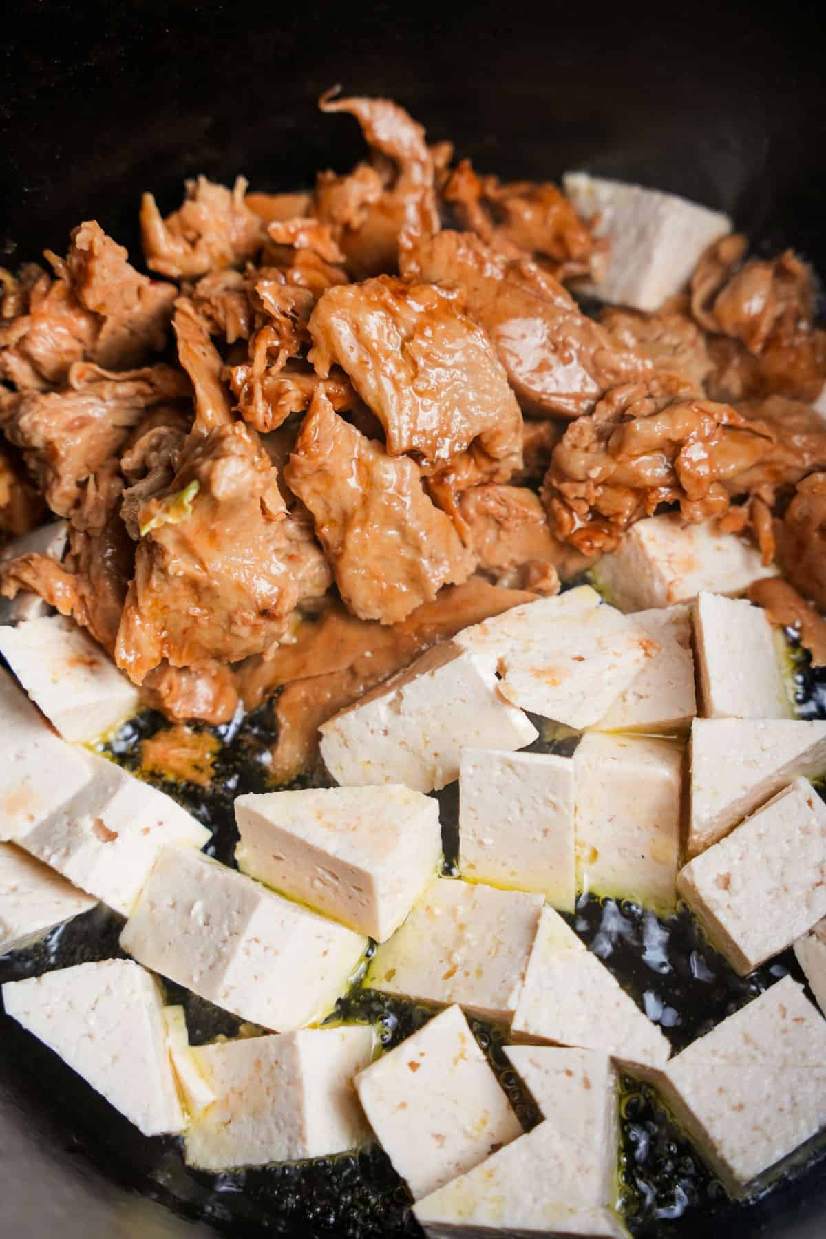 Triangles of tofu and torn bits of seitan added to hot oil in a black metal pan.