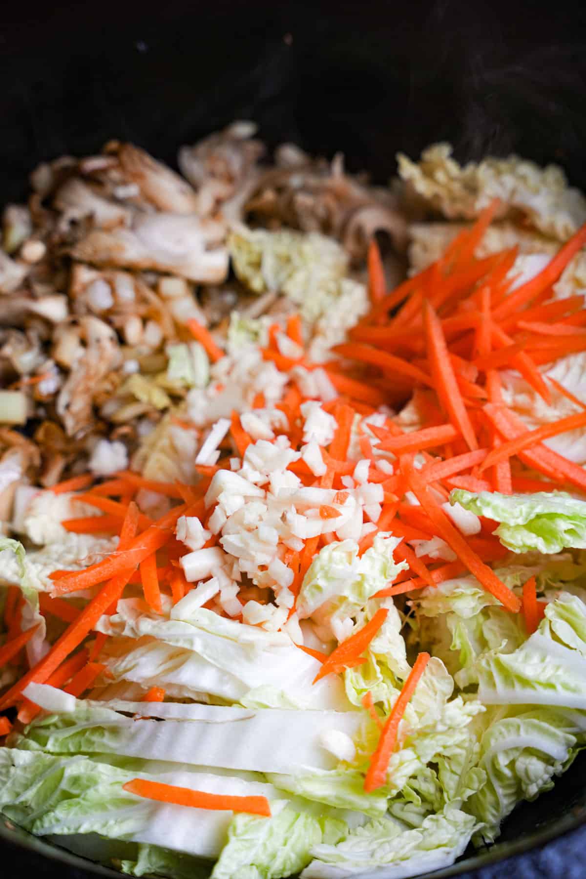 Chopped cabbage, julienne cut carrots, and minced garlic are added to the black metal pan with cooking mushrooms.