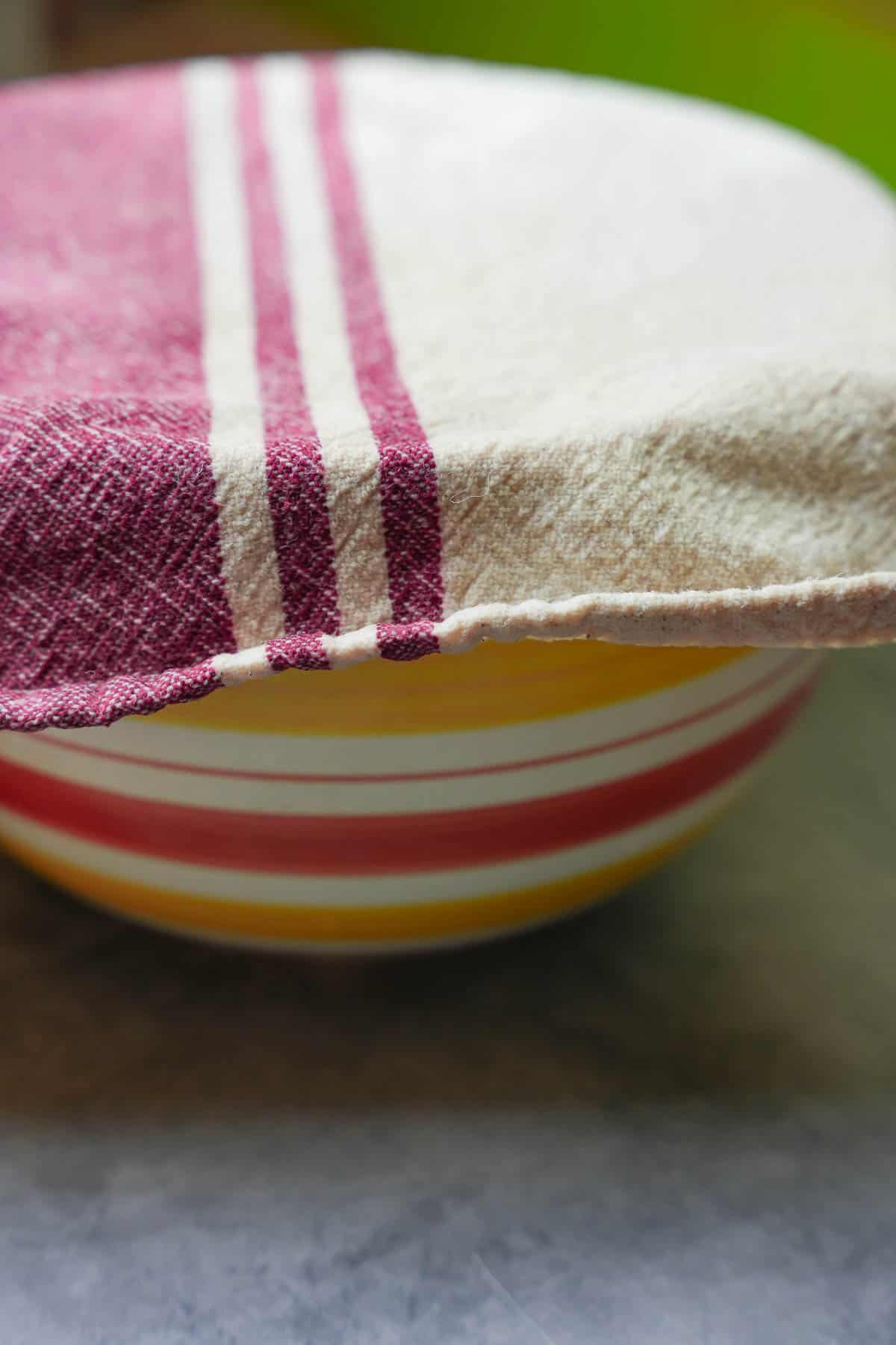 The mixed dough in a ceramic bowl is covered with a clean kitchen towel to rest and double in size.
