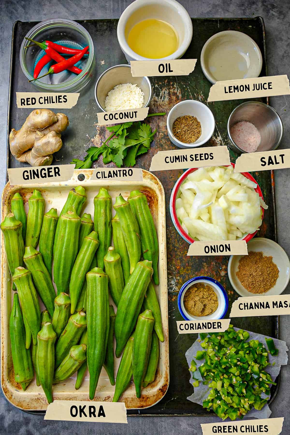 Ingredients for bhindi are measured out and labeled on top of a scratched black metal tray.
