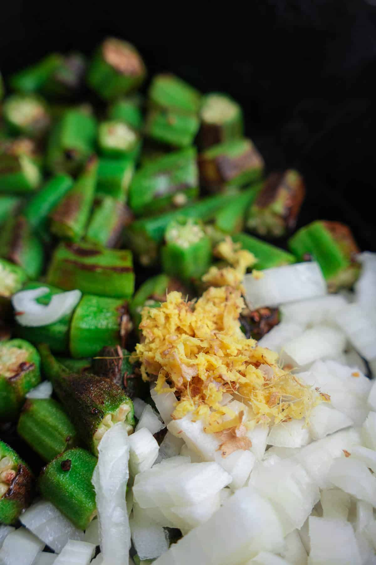 Onions and ginger are added to the sautéing okra in a cast iron pan.