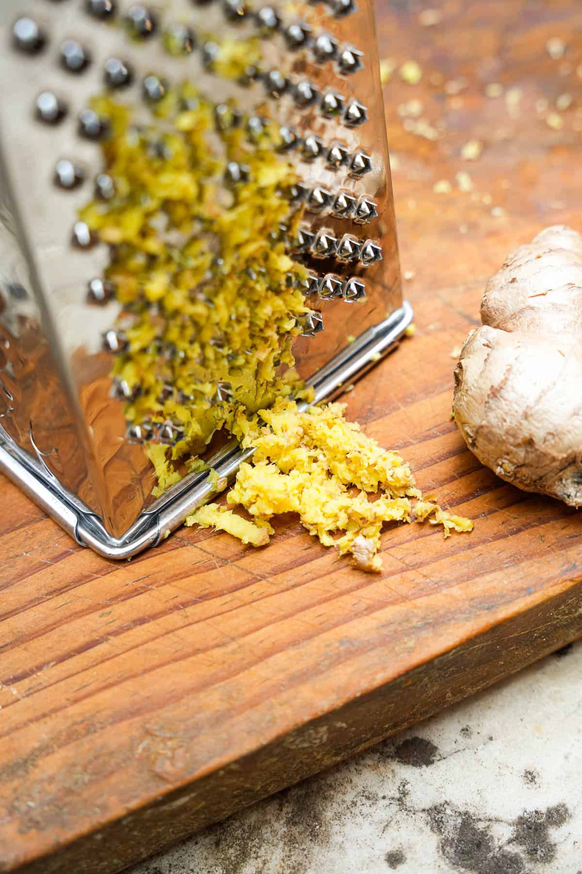 Ginger is grated using a box grater on a wooden cutting board.