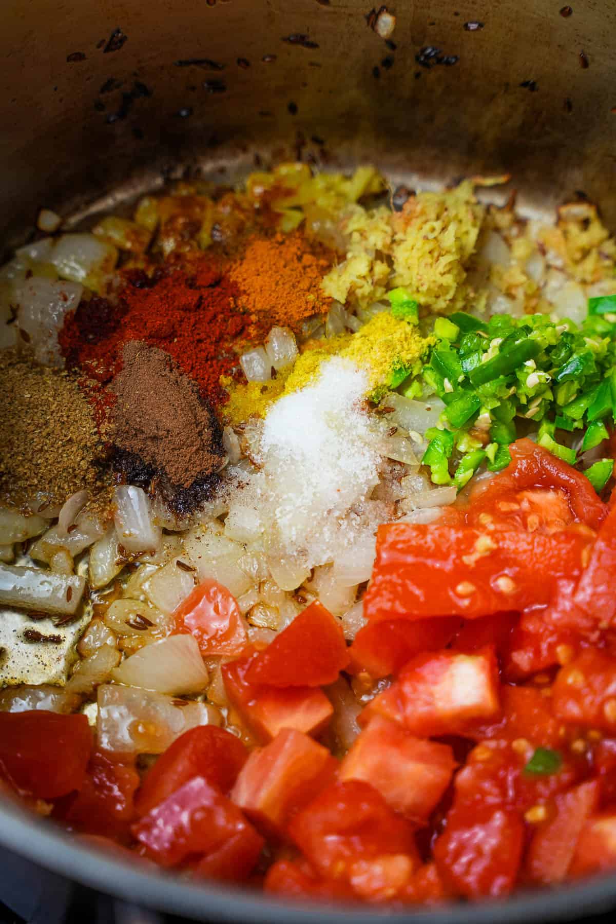 Spices, chilies, ginger, and tomatoes are added to the onions, and cumin seeds being sauteed in a stainless steel pot.