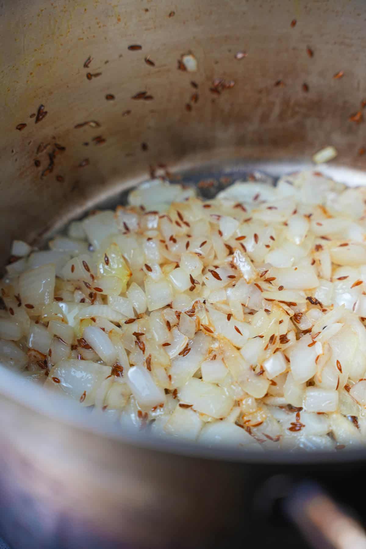 Onion sauteing with cumin seeds in a stainless steel pot.