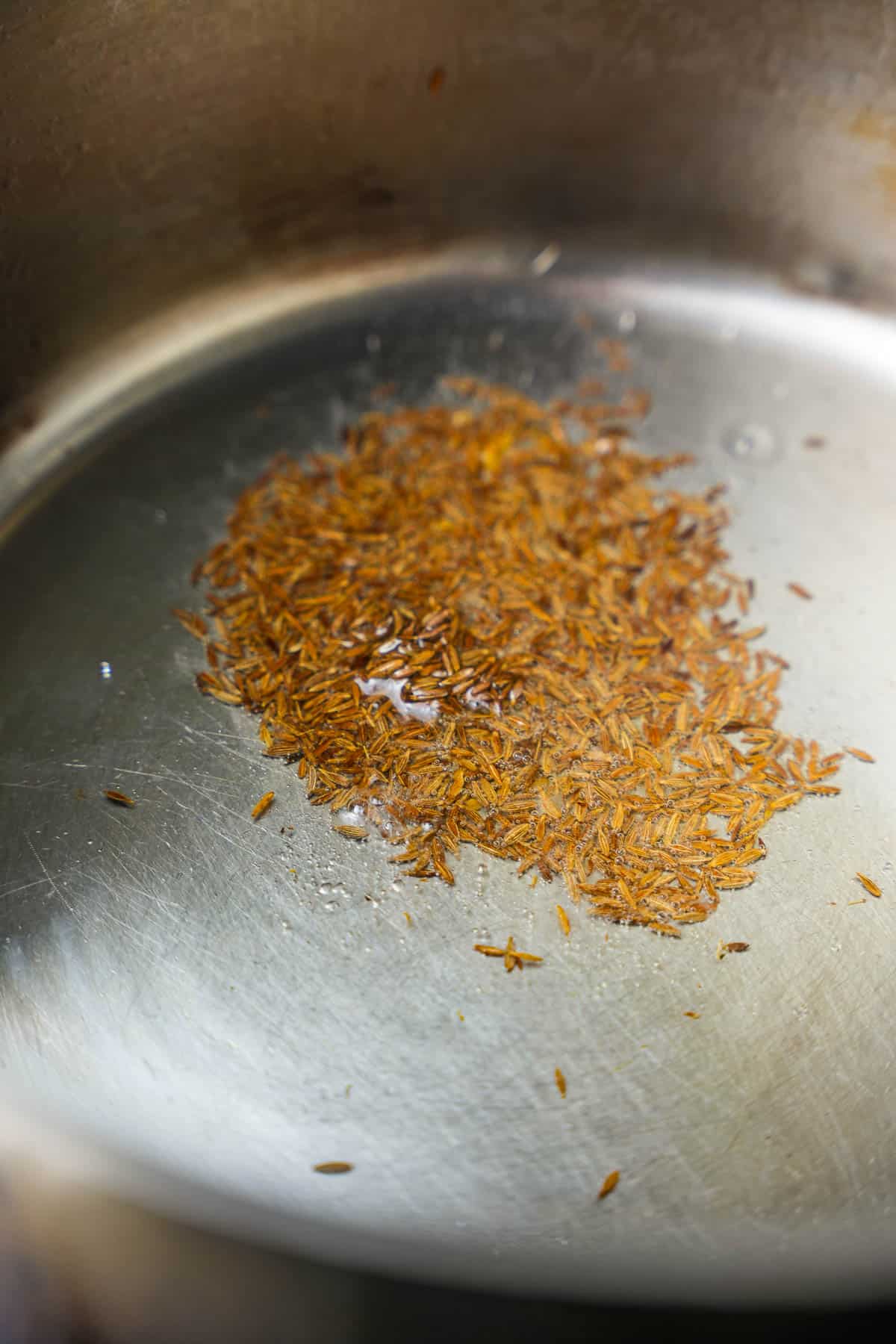Cumin seeds frying in hot oil in a stainless steel pot.