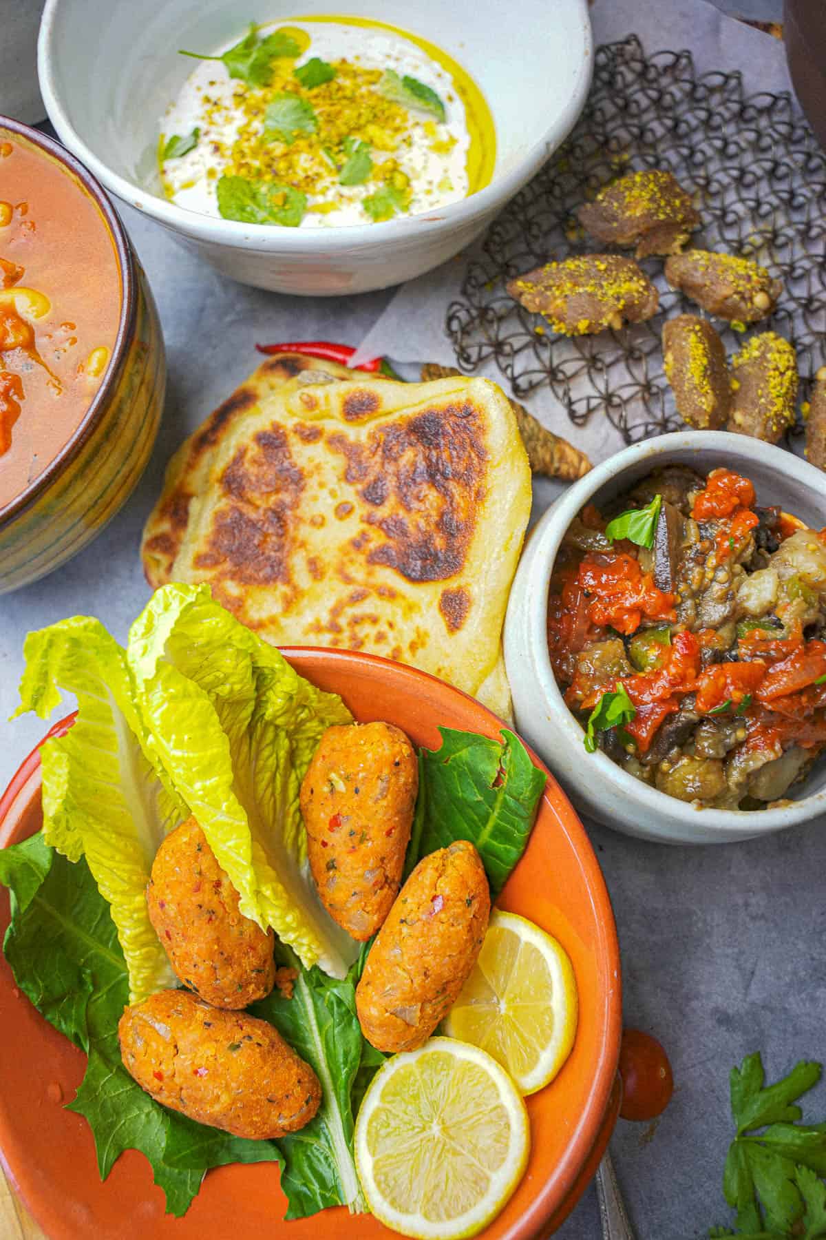 Kofte in an orange bowl with lettuce leave and lemon slices. Servings of labneh, msemen, irmik helvasi, loubia, and turkish Shakshuka are on the side.