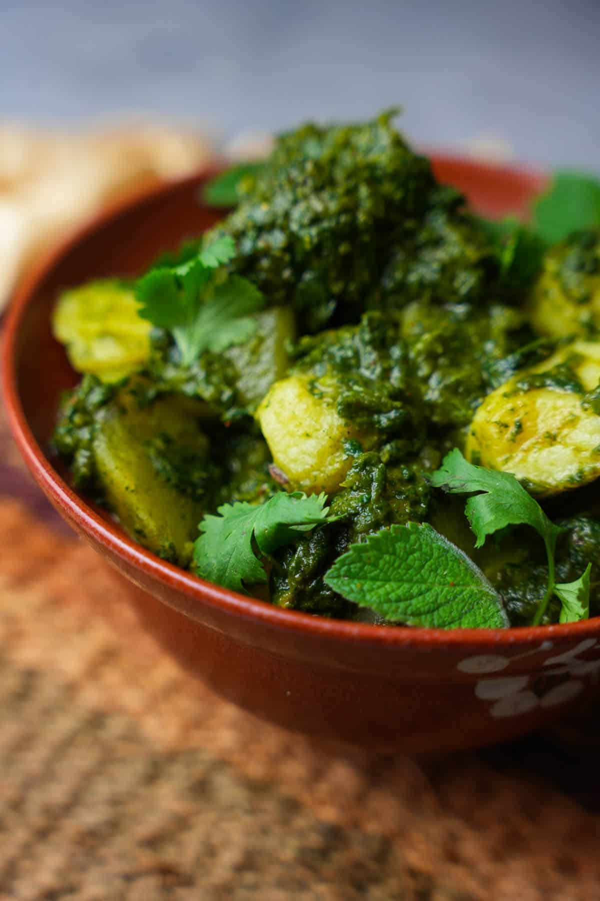 Saag aloo garnished with cilantro and mint leaves in a red ceramic bowl on a woven mat.