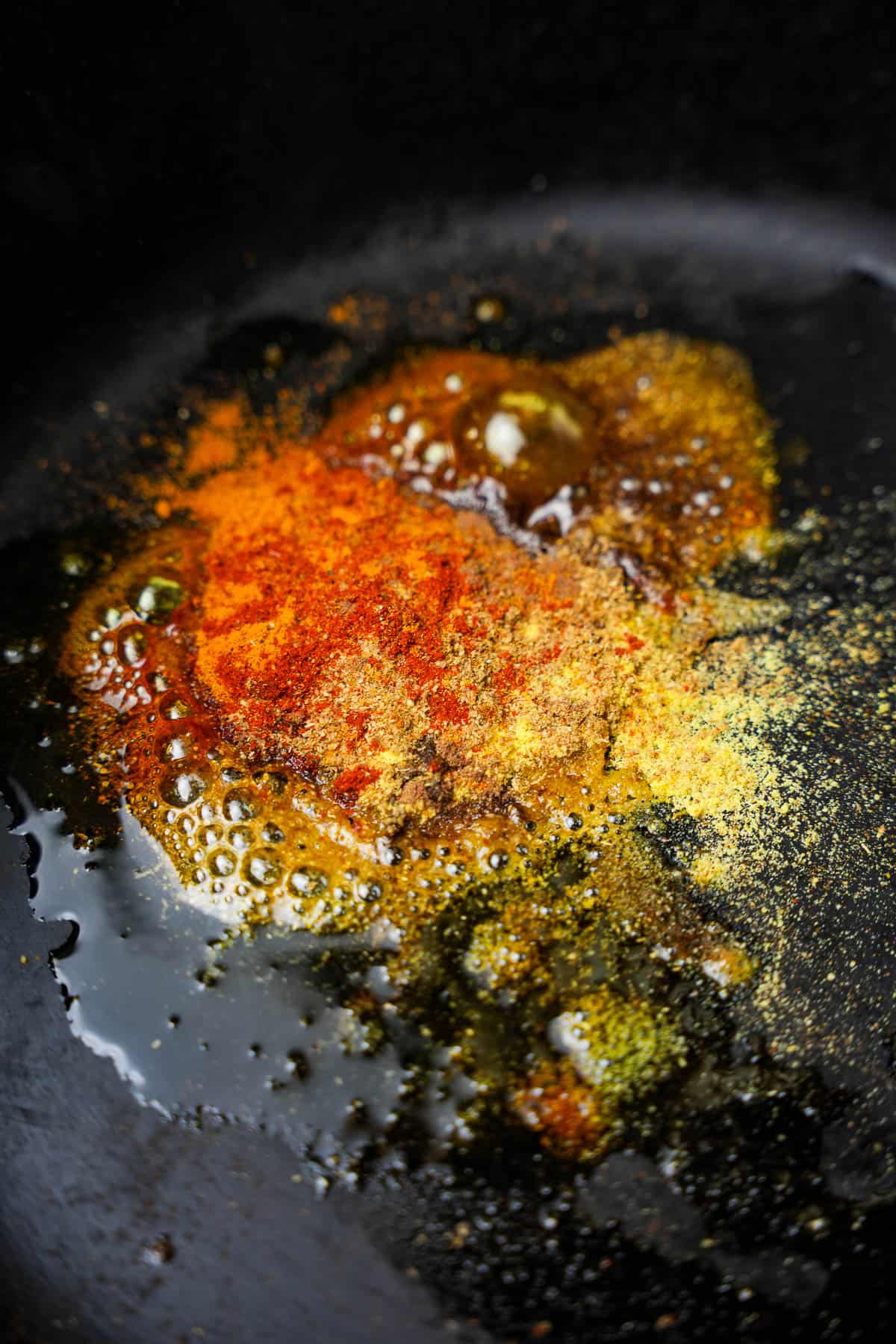 Spices are lightly fried in hot oil in a dark-colored pan.