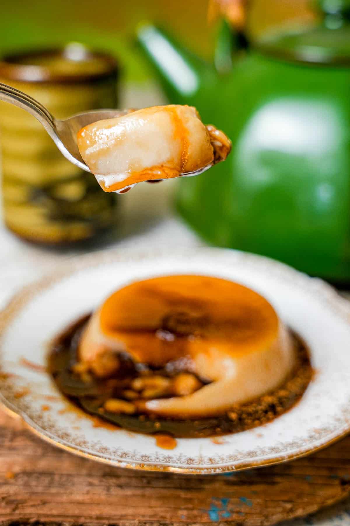 A spoonful of banh flan dripping with caramel is held over a plate with flan. A tea pot and glass can be seen in the background.