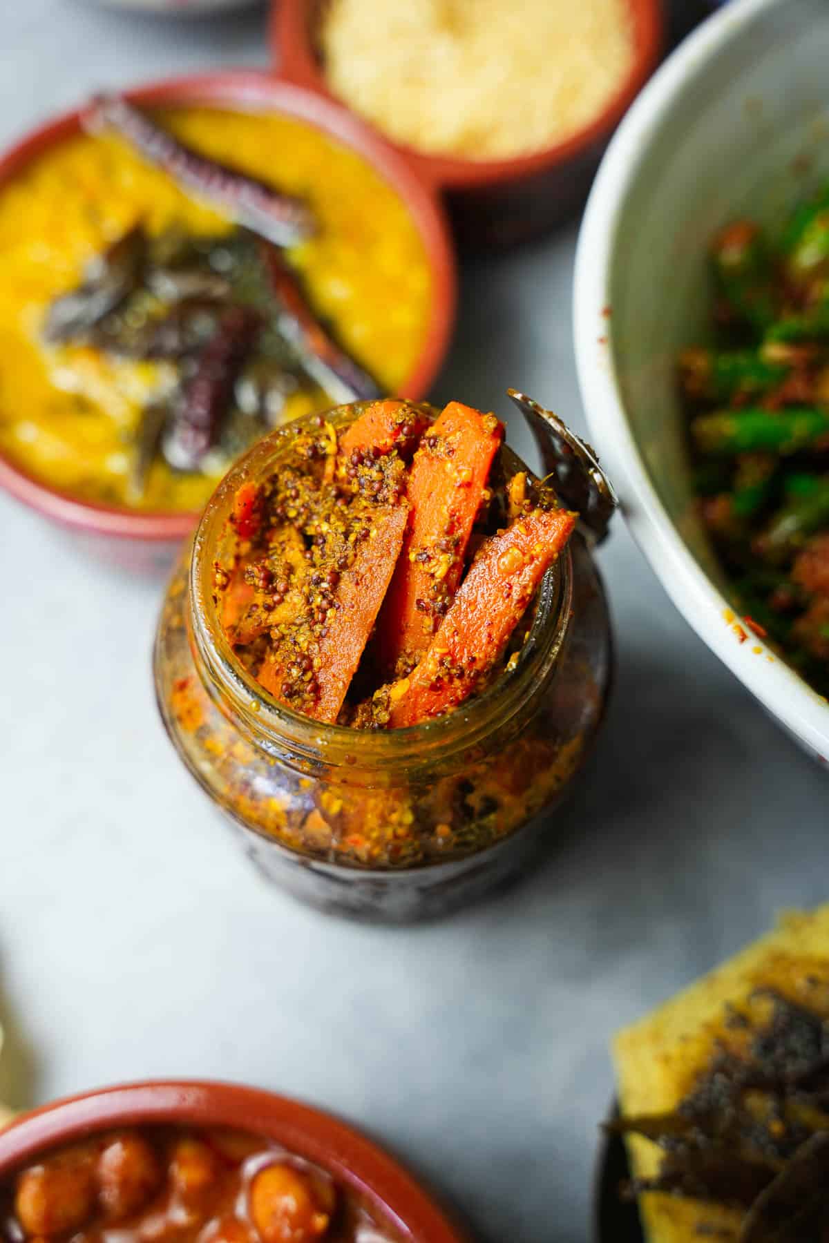 carrot pickle in glass jar surrounded by Indian dishes.