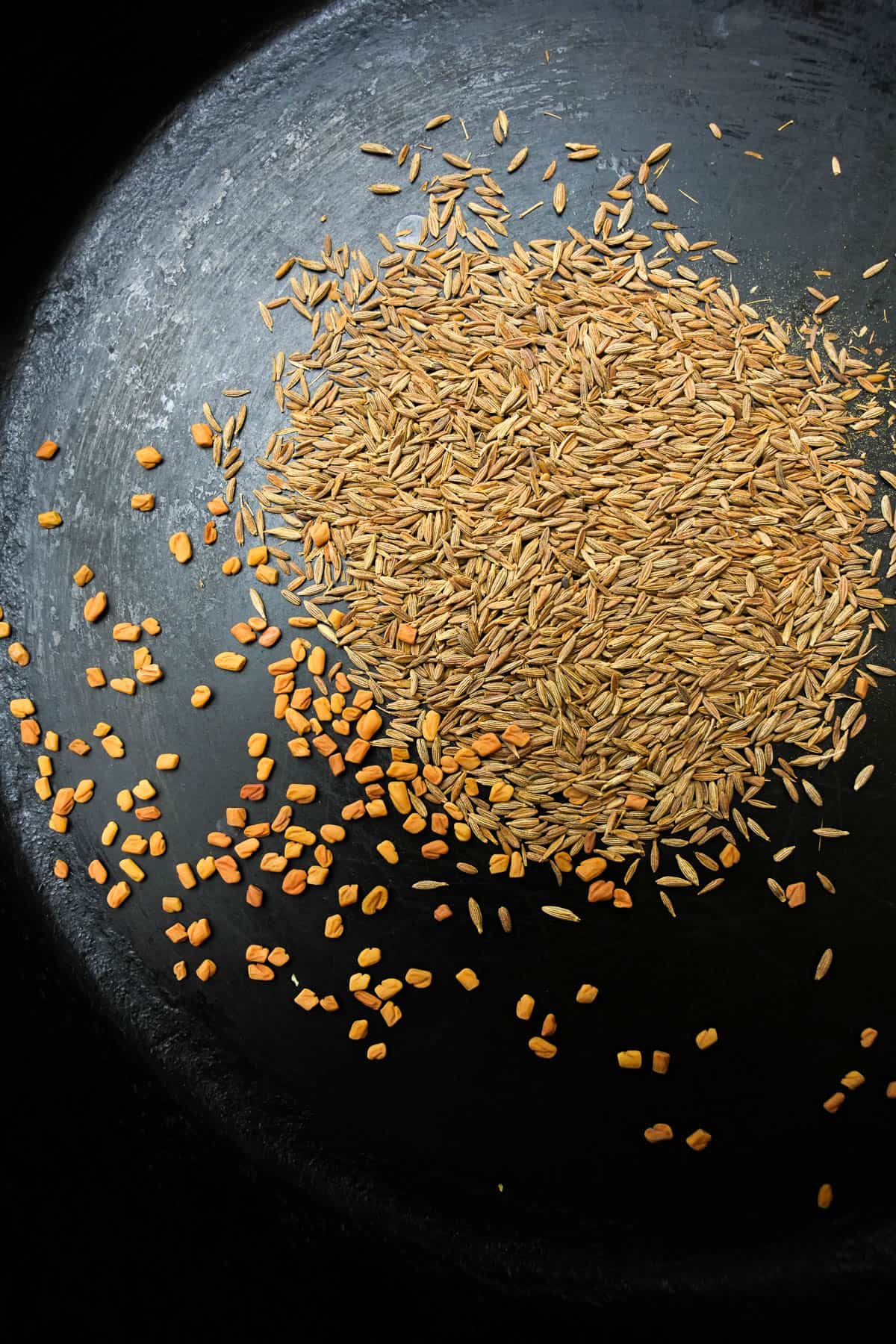 Cumin and fenugreek seeds toasting in a cast iron skillet.