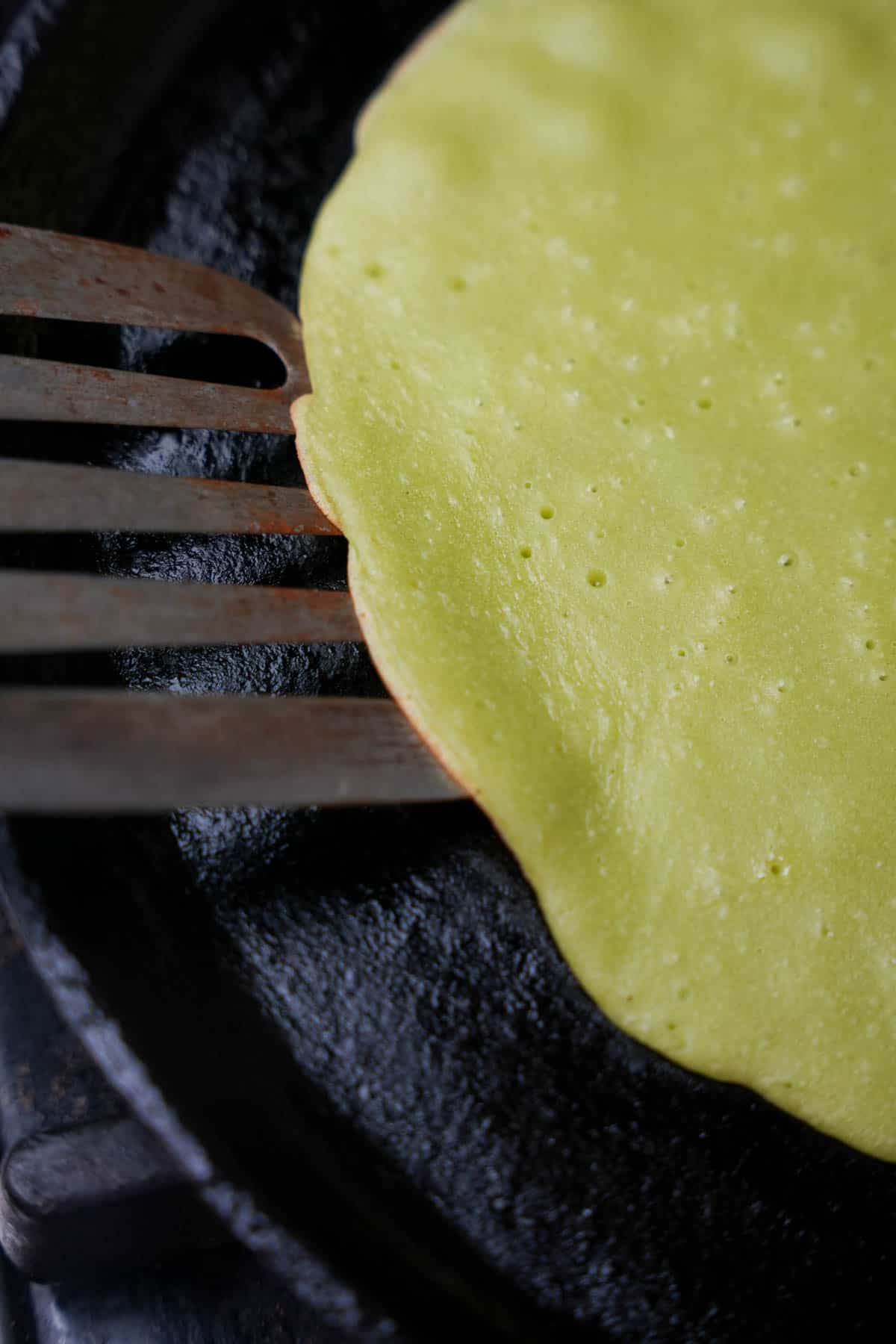 a crepe being flipped with a metal spatula in a cast iron pan.