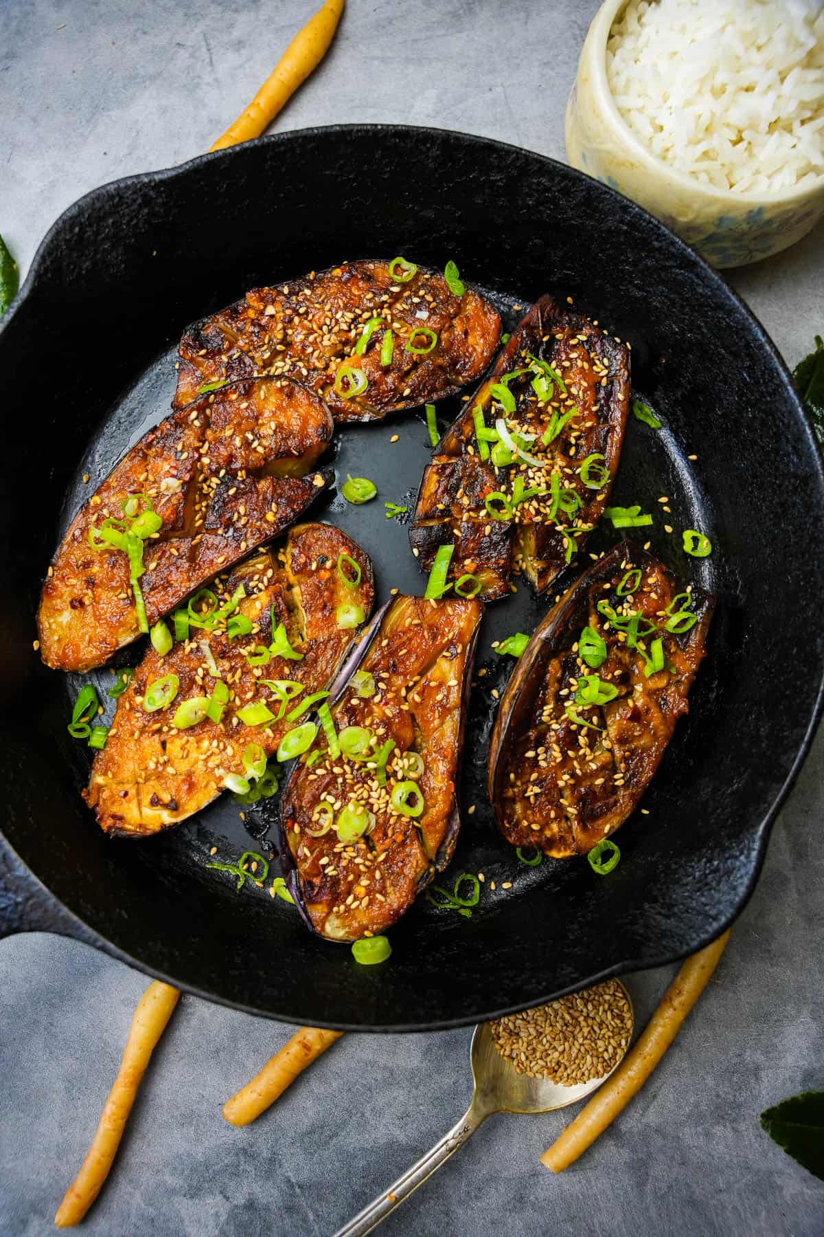Miso glazed eggplant in a skillet with rice and green onions. Yamagobo and sesame seeds are on the counter top.
