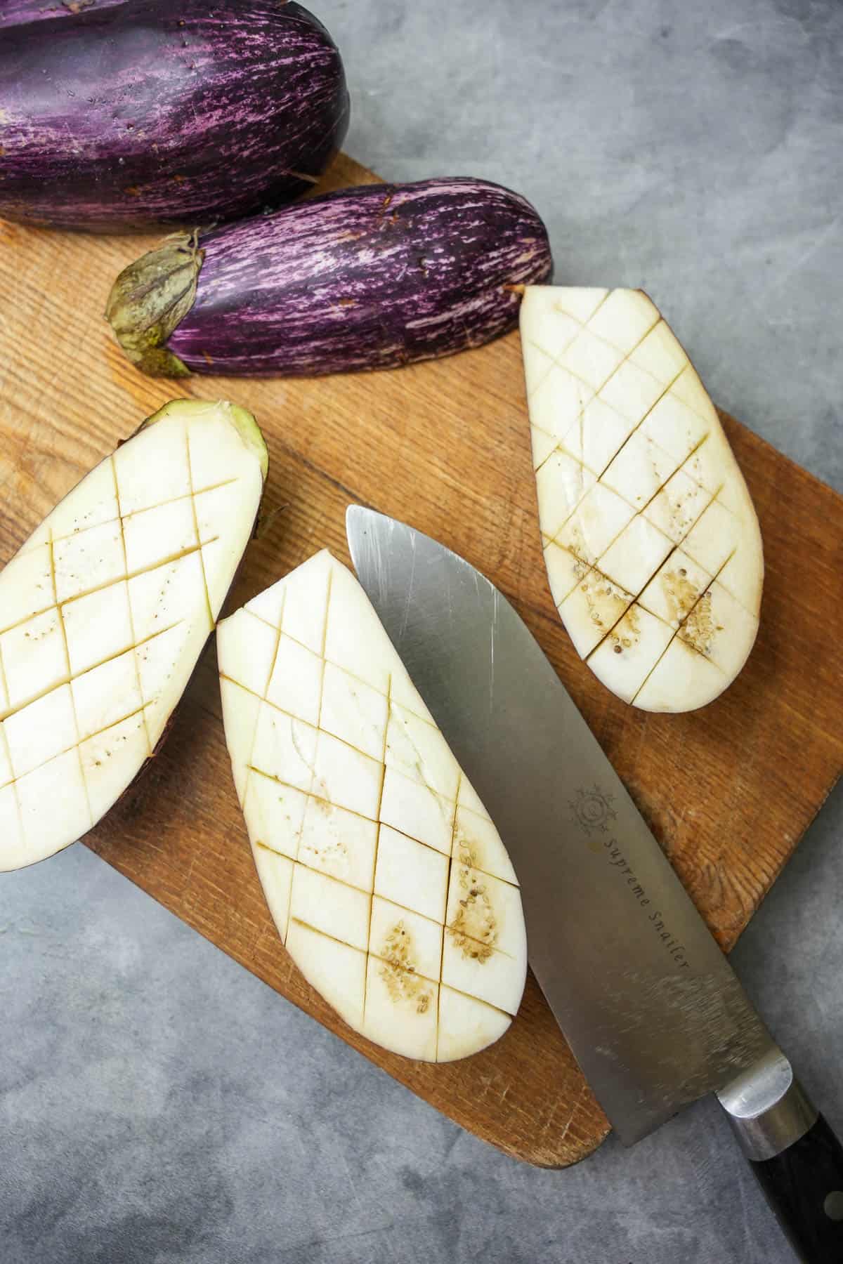the surface of eggplant is cut into a diamond pattern with a sharp knife on a wooden cutting board.