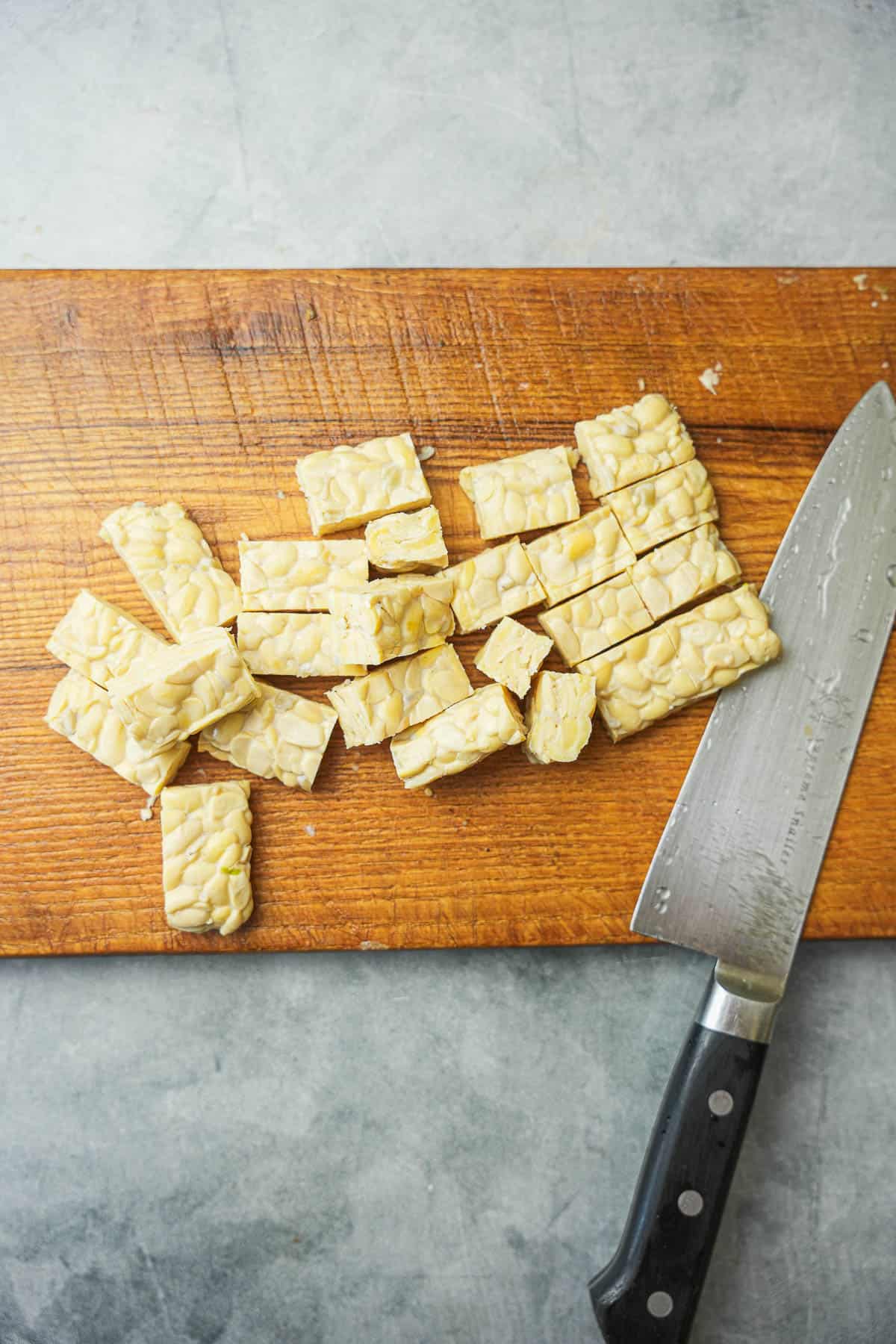 Tempeh cut into bite sized pieces on a wooden cutting board with a sharp knife.