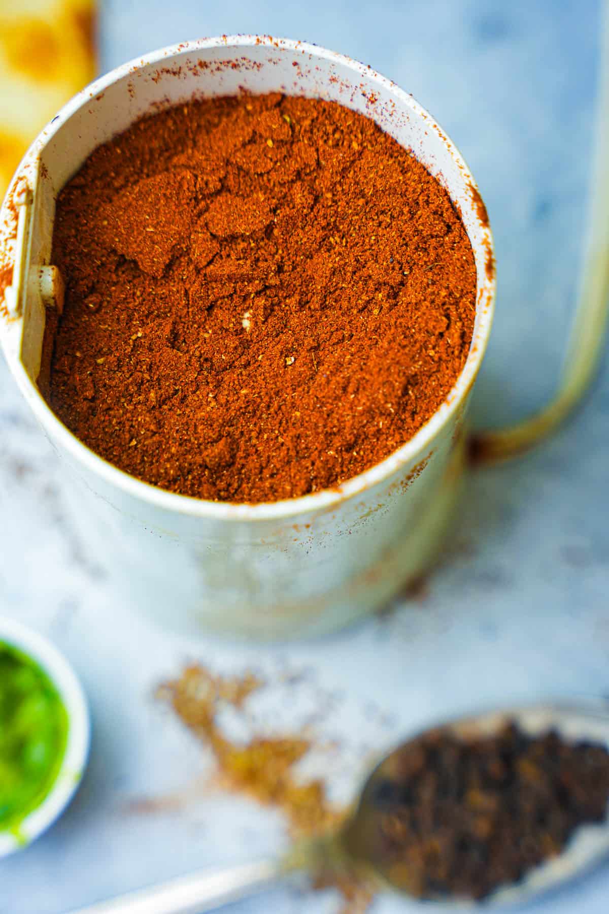 A white spice grinder filled with a mixture of spices and a spoon.