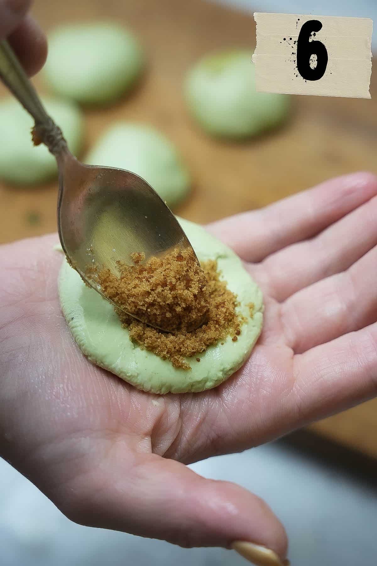 A person is holding a spoon with palm sugar, adding it to a flattened portion of pandan dough in their other hand.