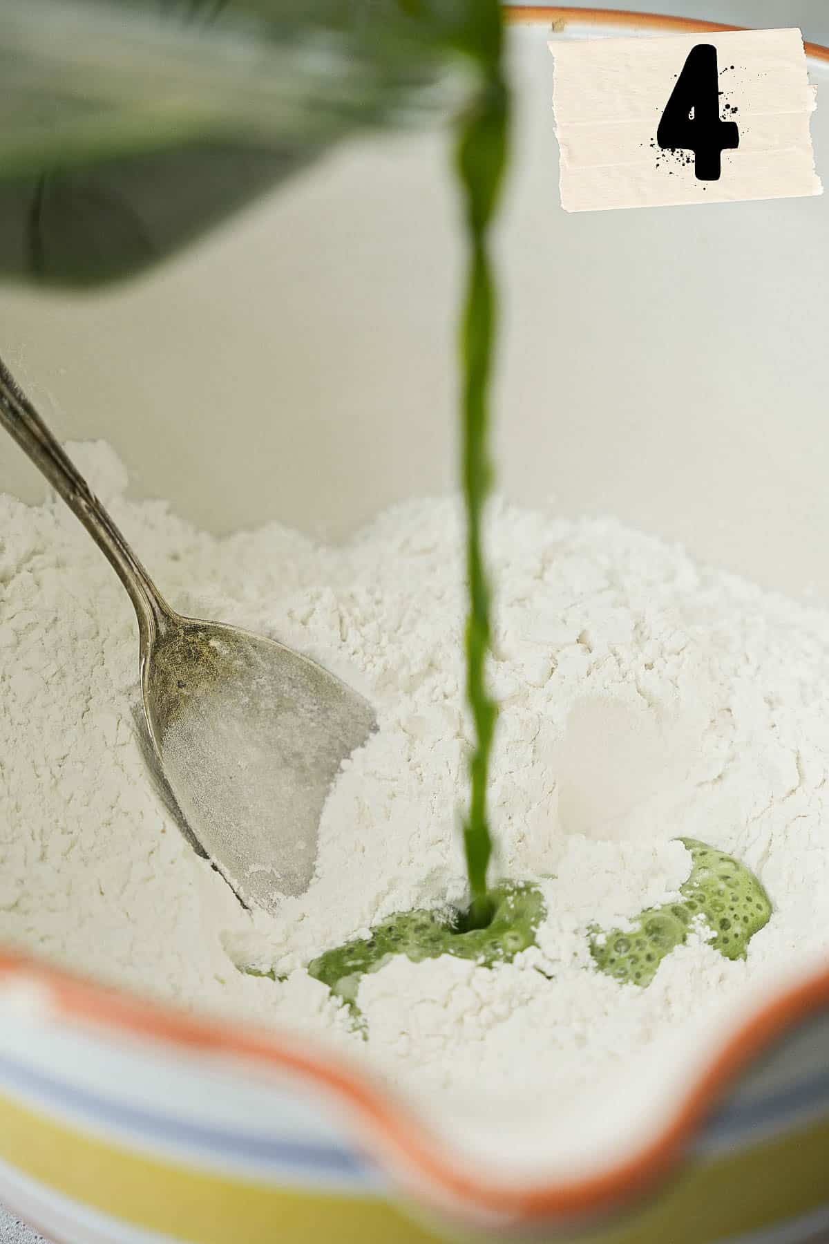 pandan juice is being poured into a bowl of flour with a metal spoon in it.