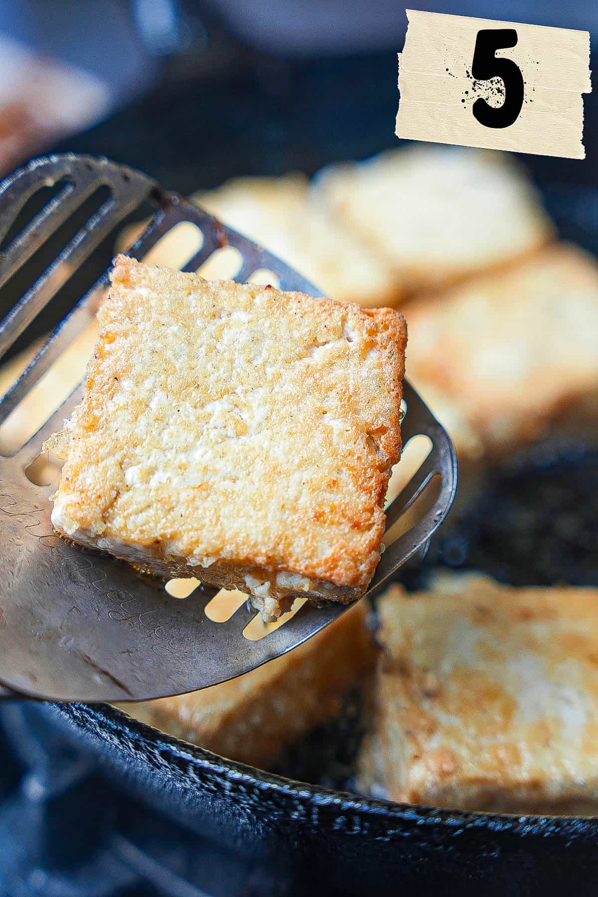 Tofu frying in a pan. A spatula removes one crispy piece.