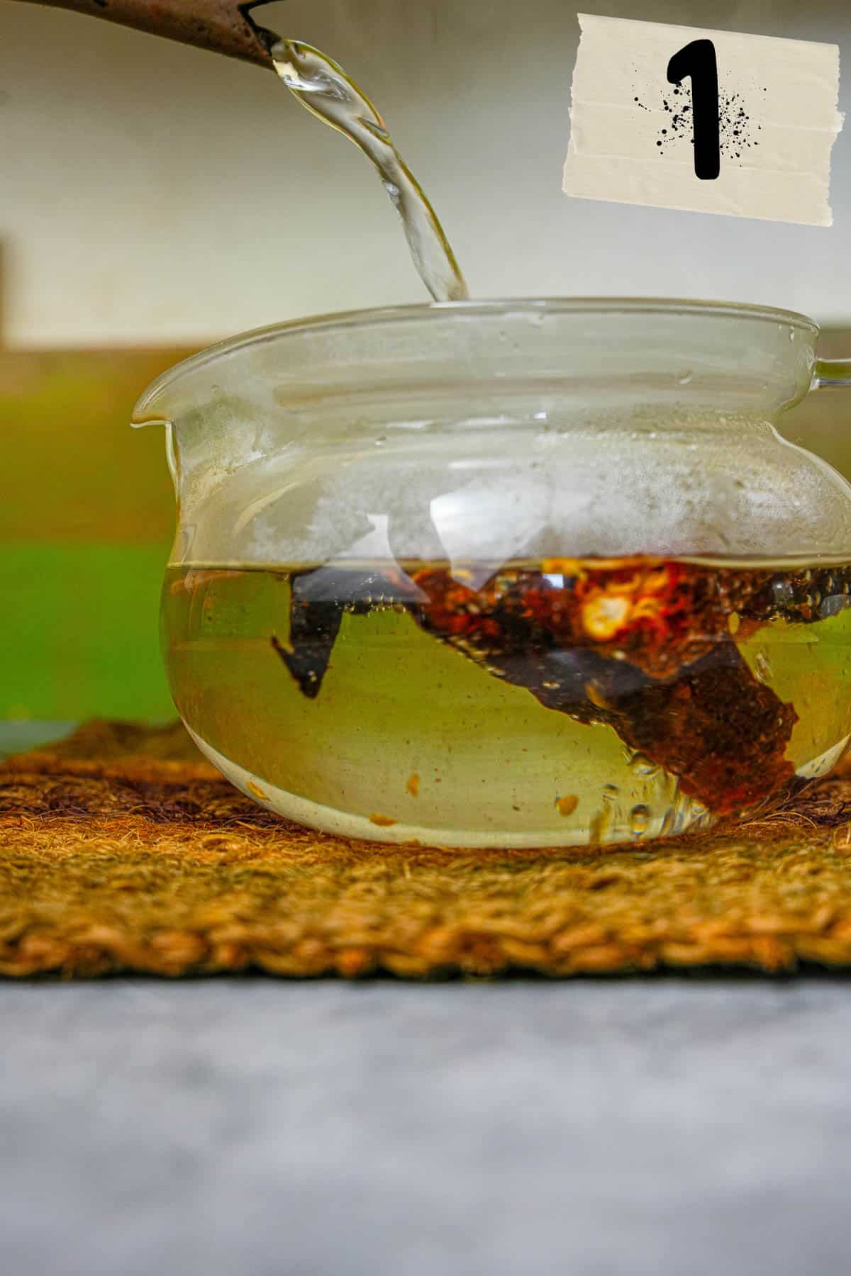 A person pouring hot water into a glass bowl with dried chilies in it.