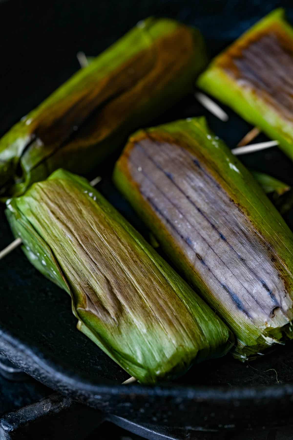 Tupig in banana leaves cooking in a skillet with toothpicks to seal them.