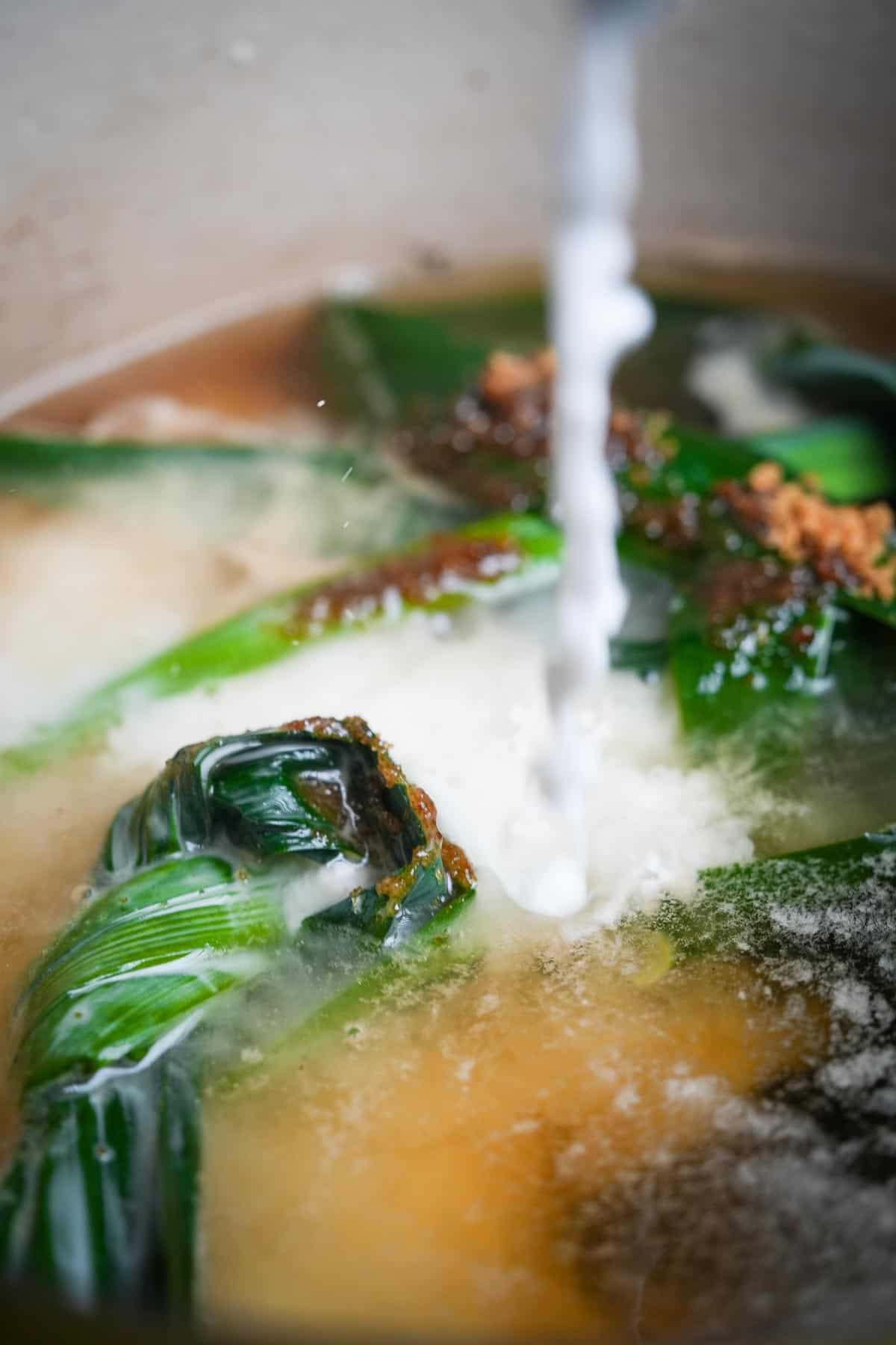 Coconut milk being poured into the pot of boiling pandan leaves.