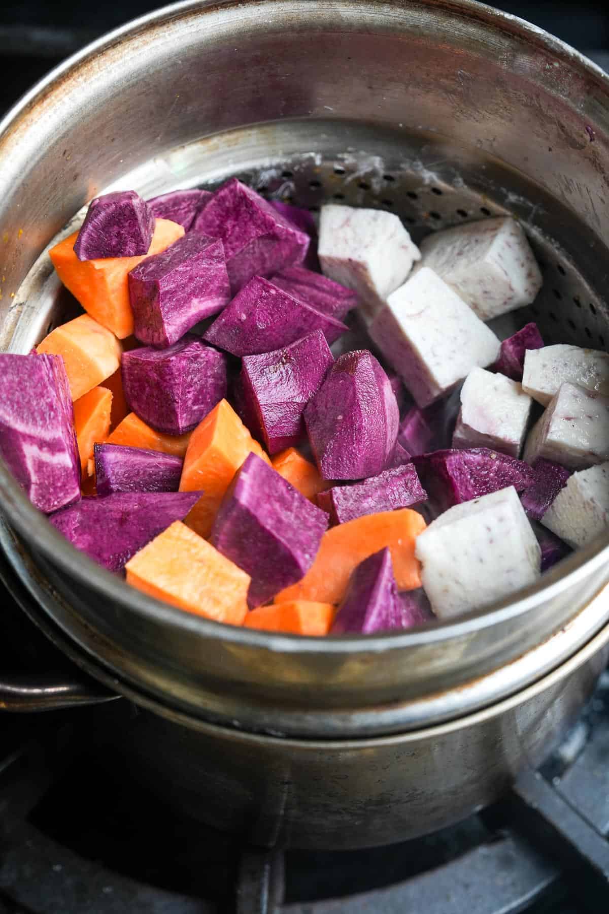 A steamer full of purple sweet potatoes, orange sweet potatoes and taro on a stove.