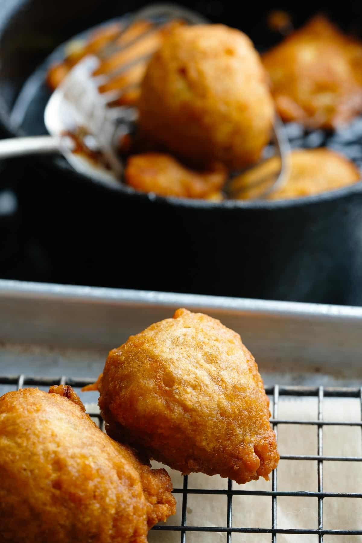 Fried banana doughnuts cooling on a wire rack next to a pan that has more cooking in it.