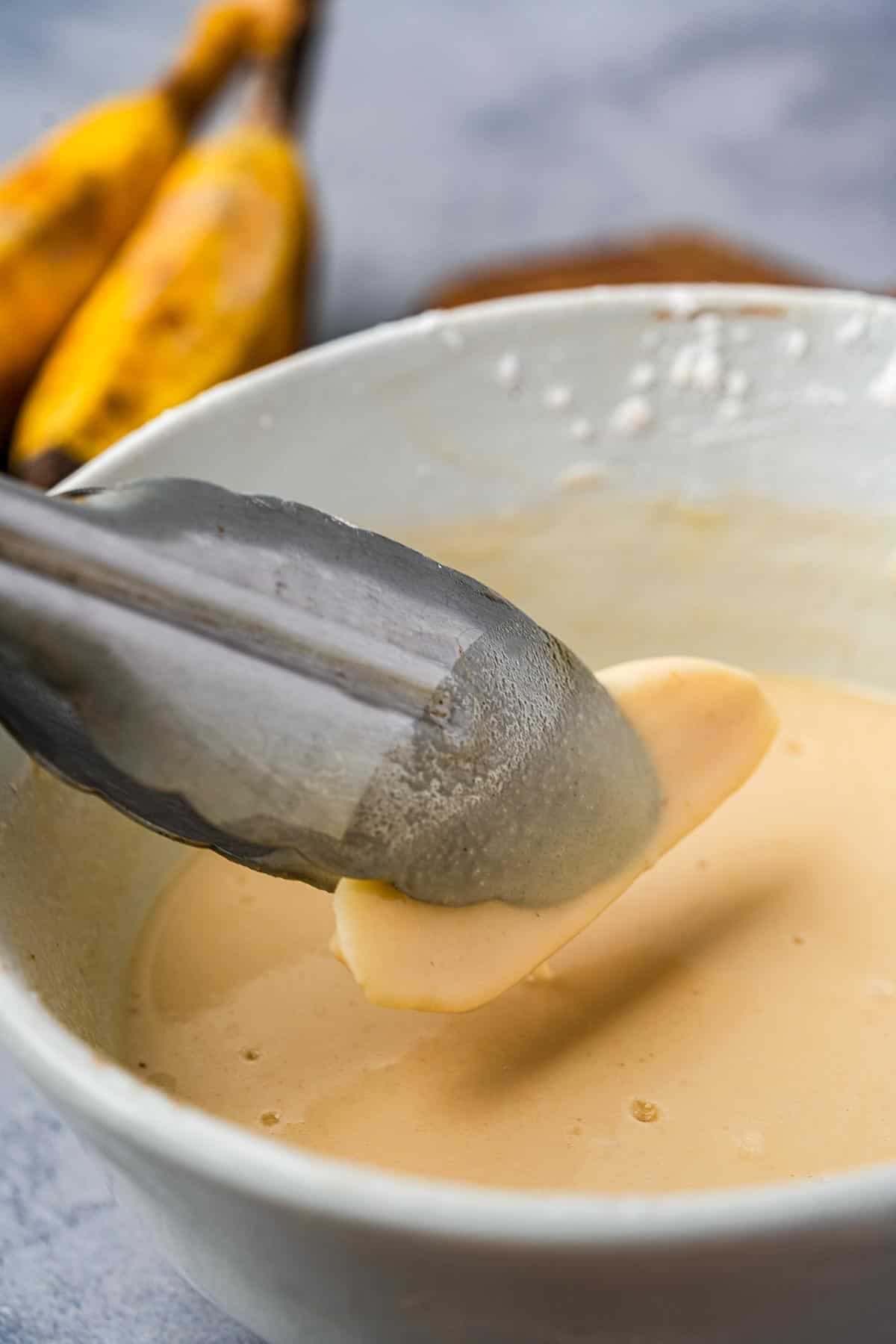 Tongs are being used to top bananas into a bowl of batter.