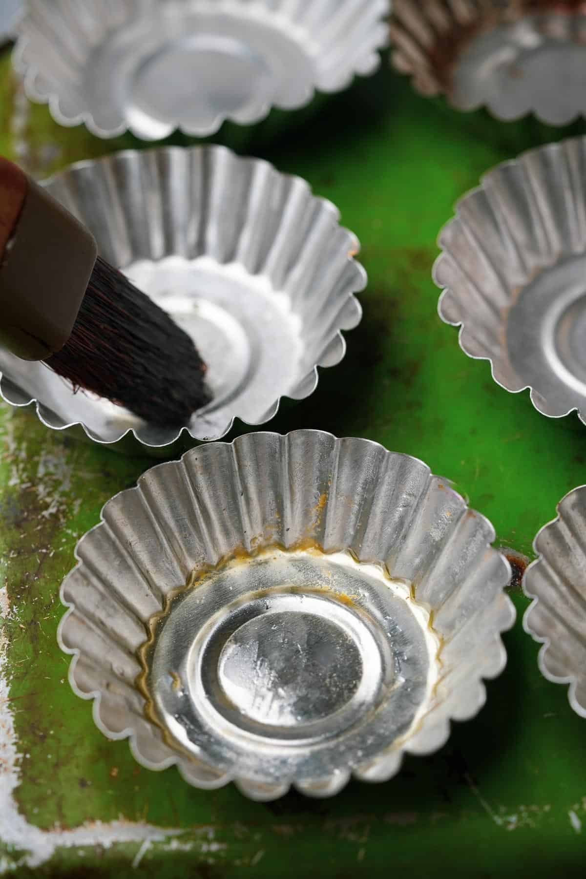 A person is using a brush to oil a set of metal cake molds.