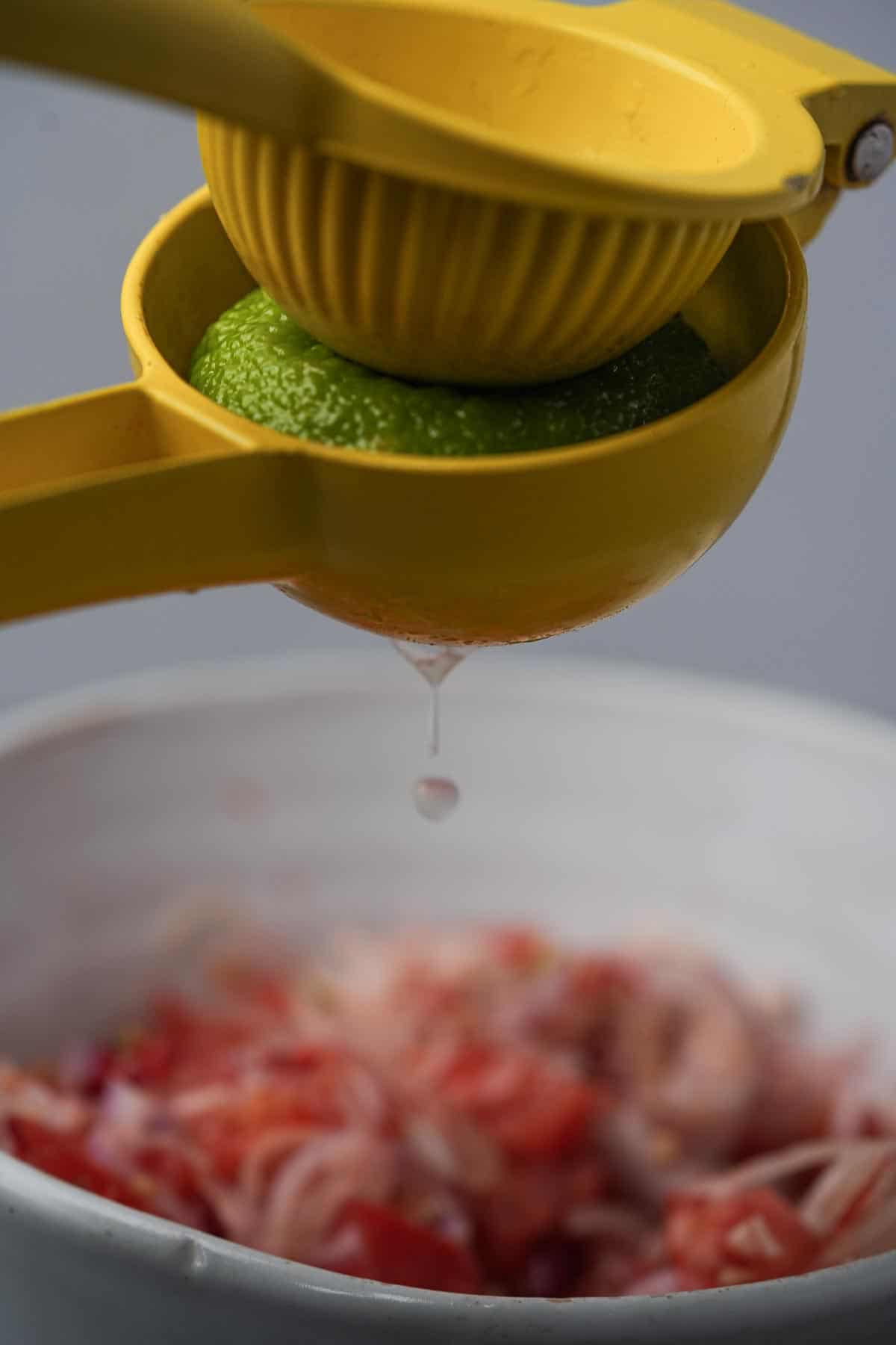 A yellow citrus squeezer is being used to juice lime into a bowl of shallots and tomatoes.
