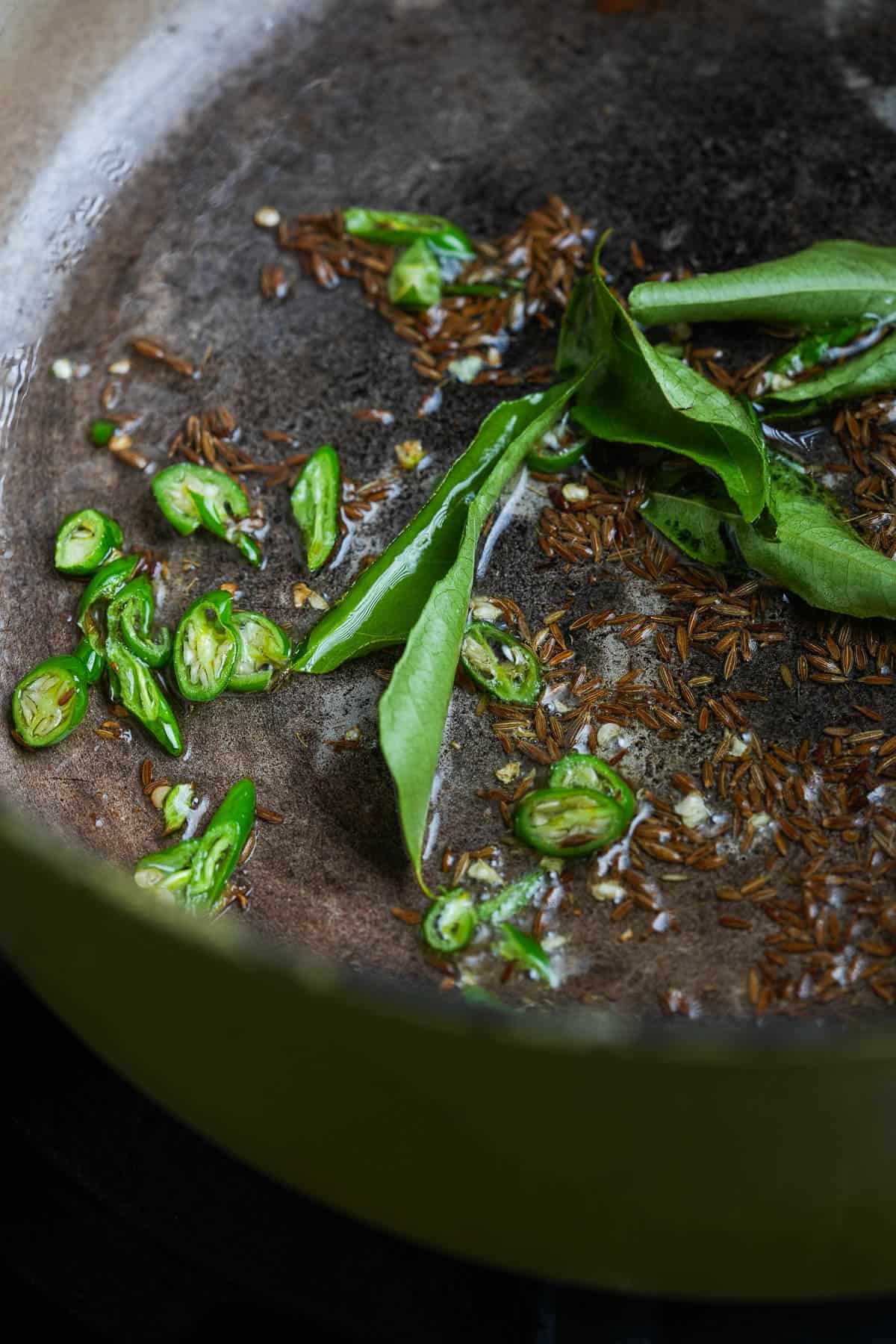 A frying pan with green curry leaves, chilies, and cumin seeds frying in it.