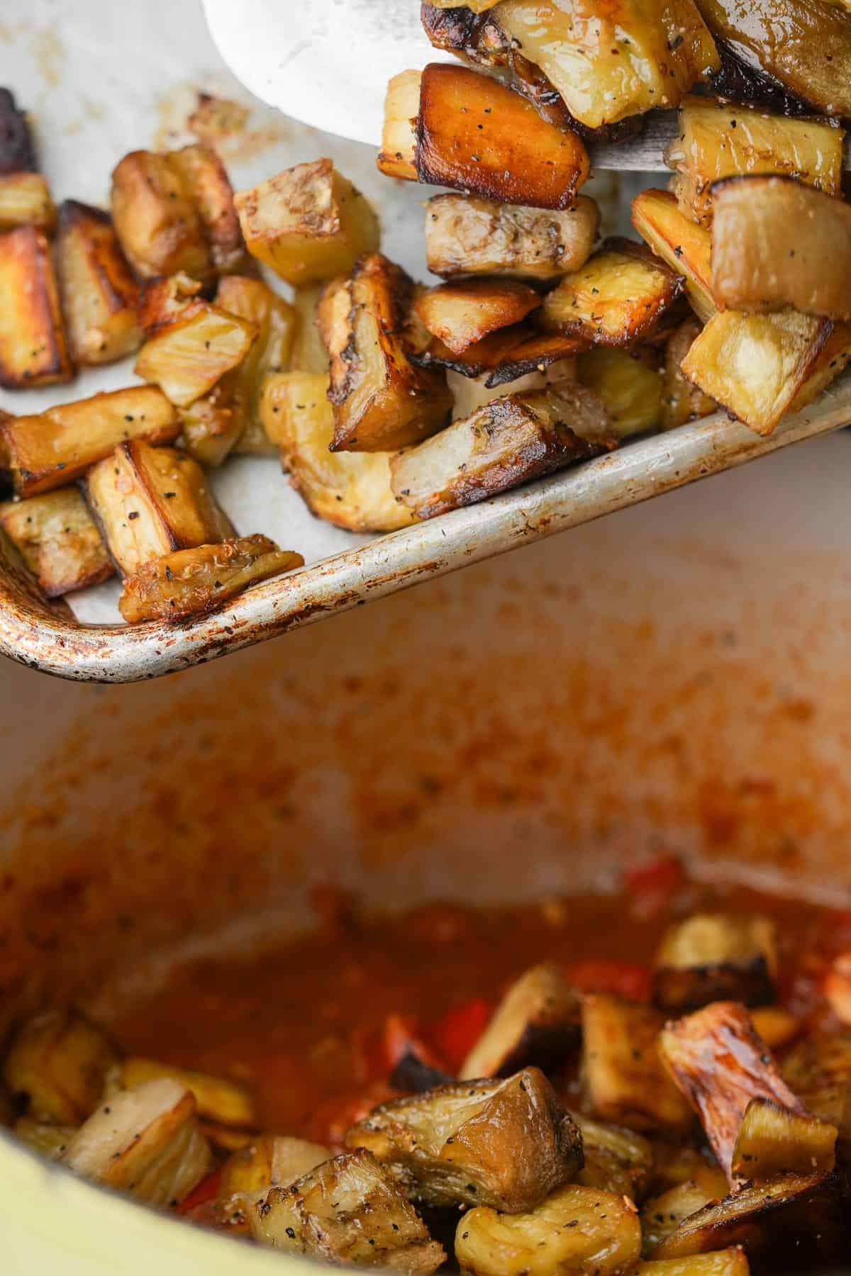 A tray of roasted eggplant is added to a pot of cooking red sauce.
