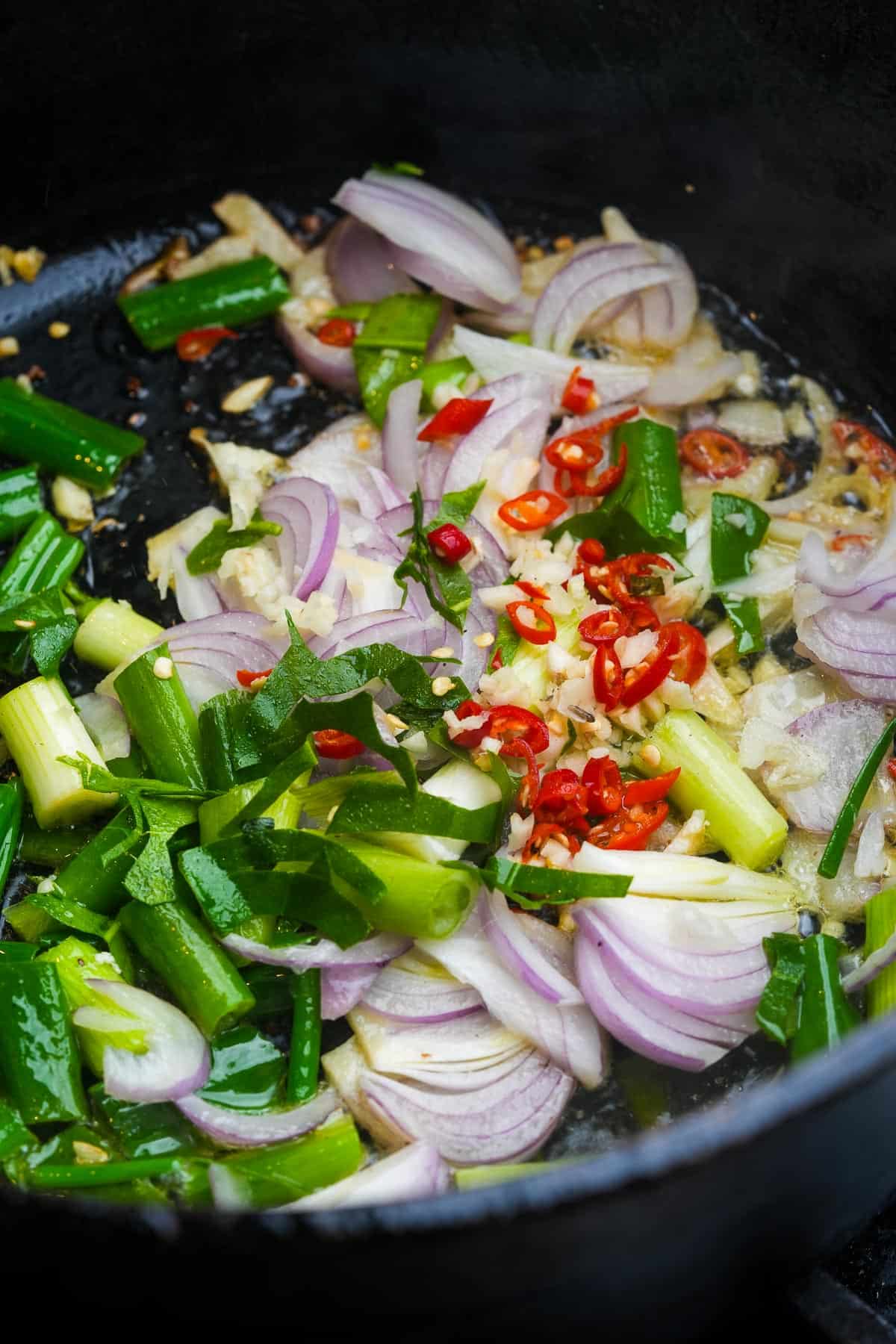 A frying pan with shallots, lime leaves, scallions and peppers in it.