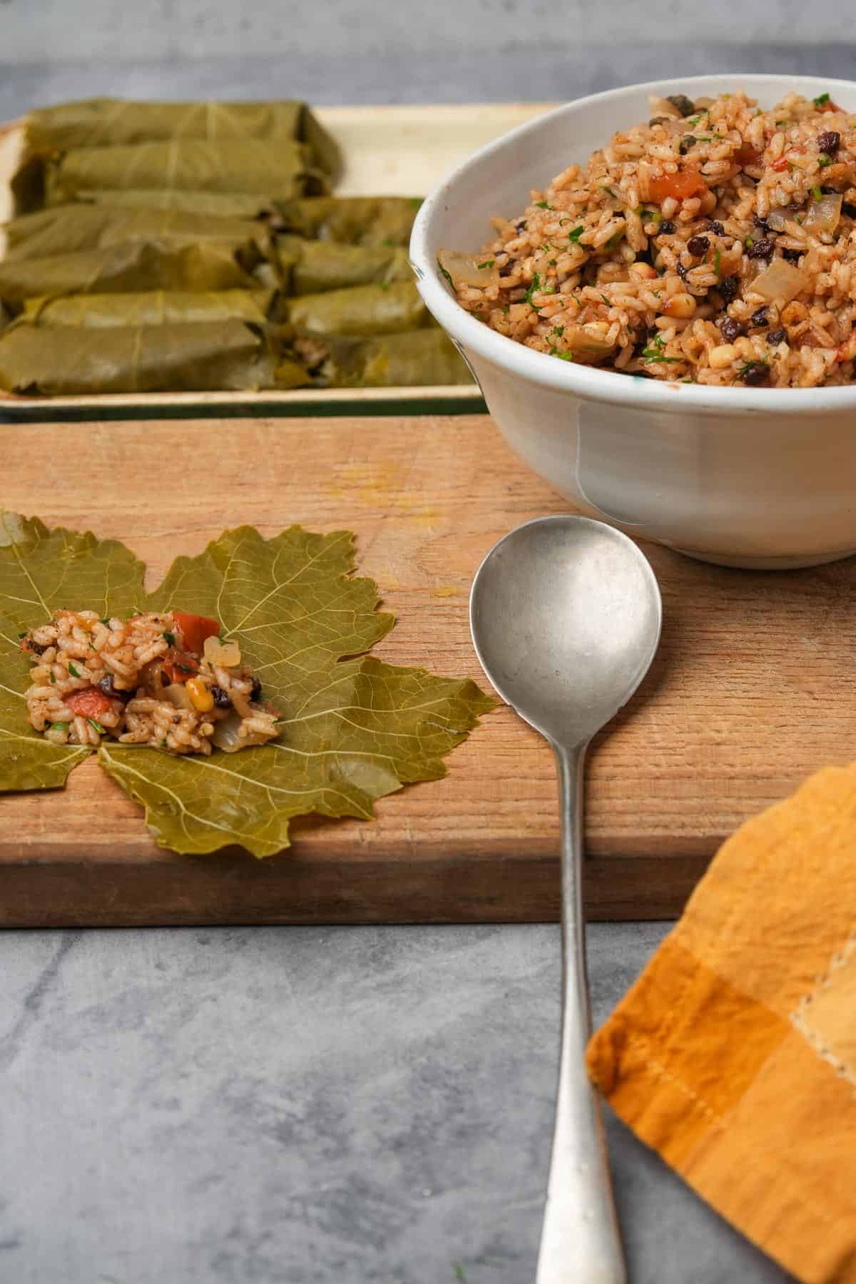 A bowl of rice filling and some being placed in the center of a grape leaf. Wrapped stuffed grape leaves behind on a cutting board.